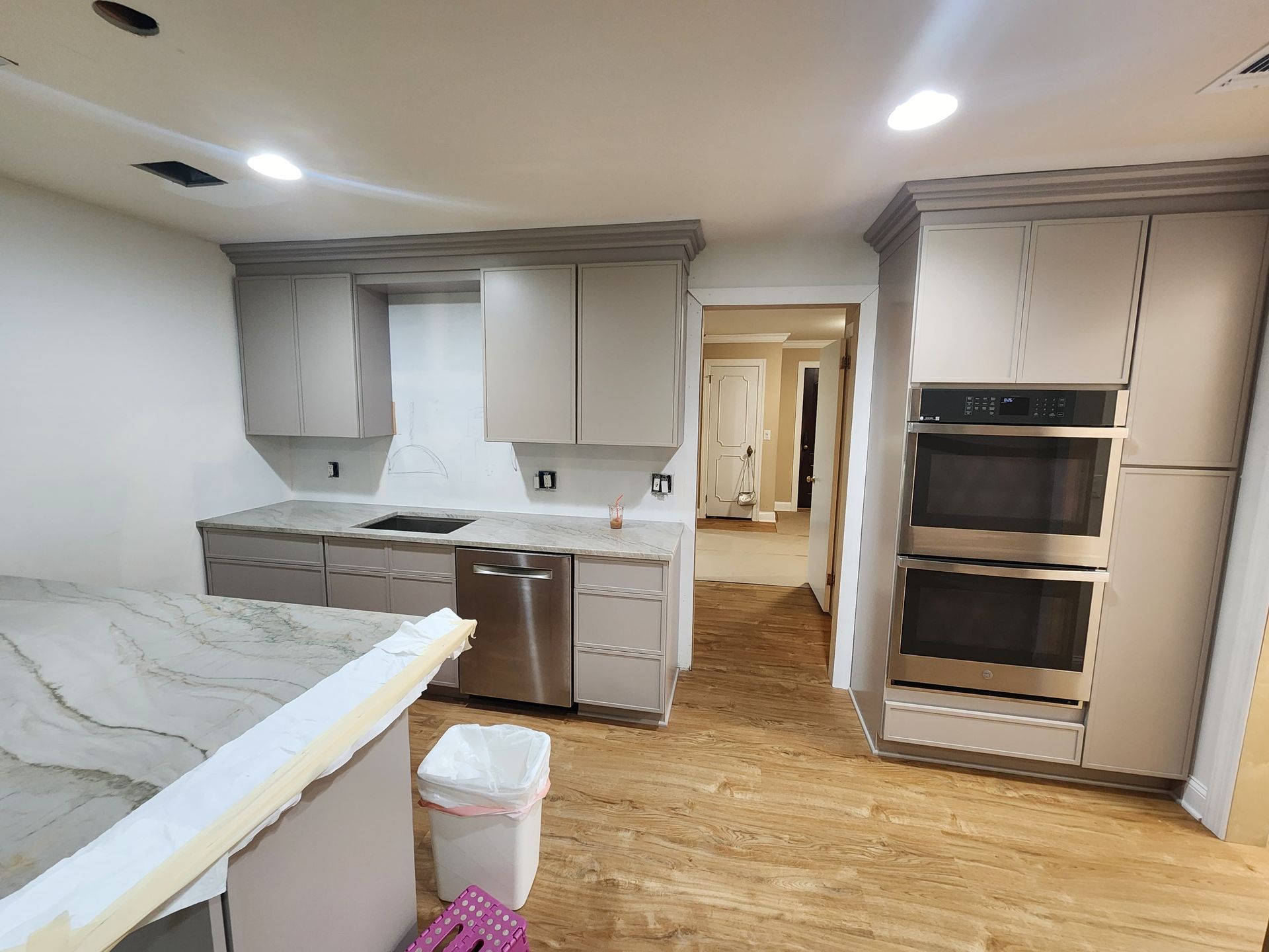 A renovated kitchen featuring light taupe cabinets, a stainless steel double oven, a dishwasher, and stone countertops.