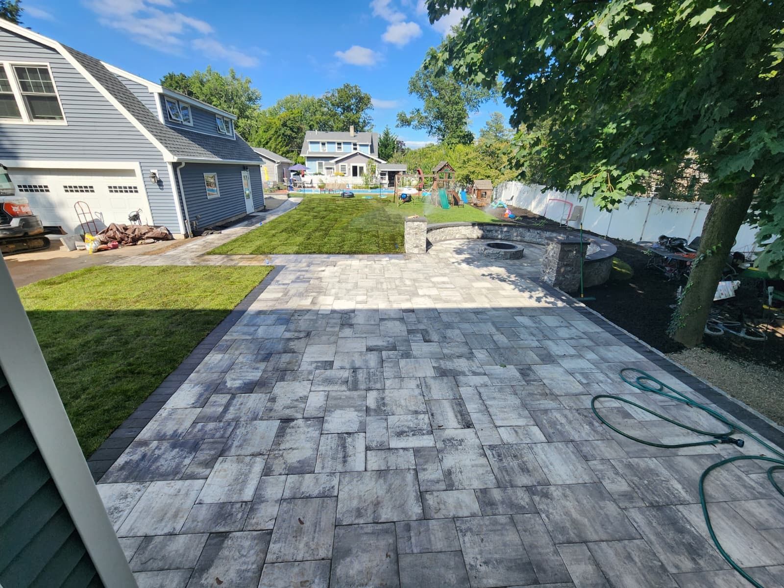 A backyard featuring a newly laid grey stone patio, a small lawn, and a fire pit area next to a light blue house.