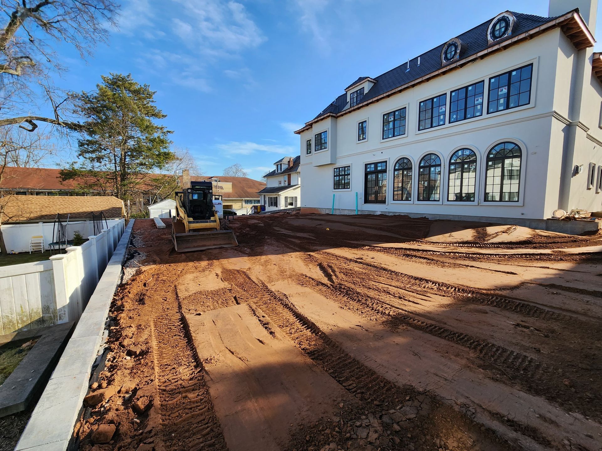 A construction site with a large white house and a parked excavator on uneven, graded dirt under a clear blue sky.