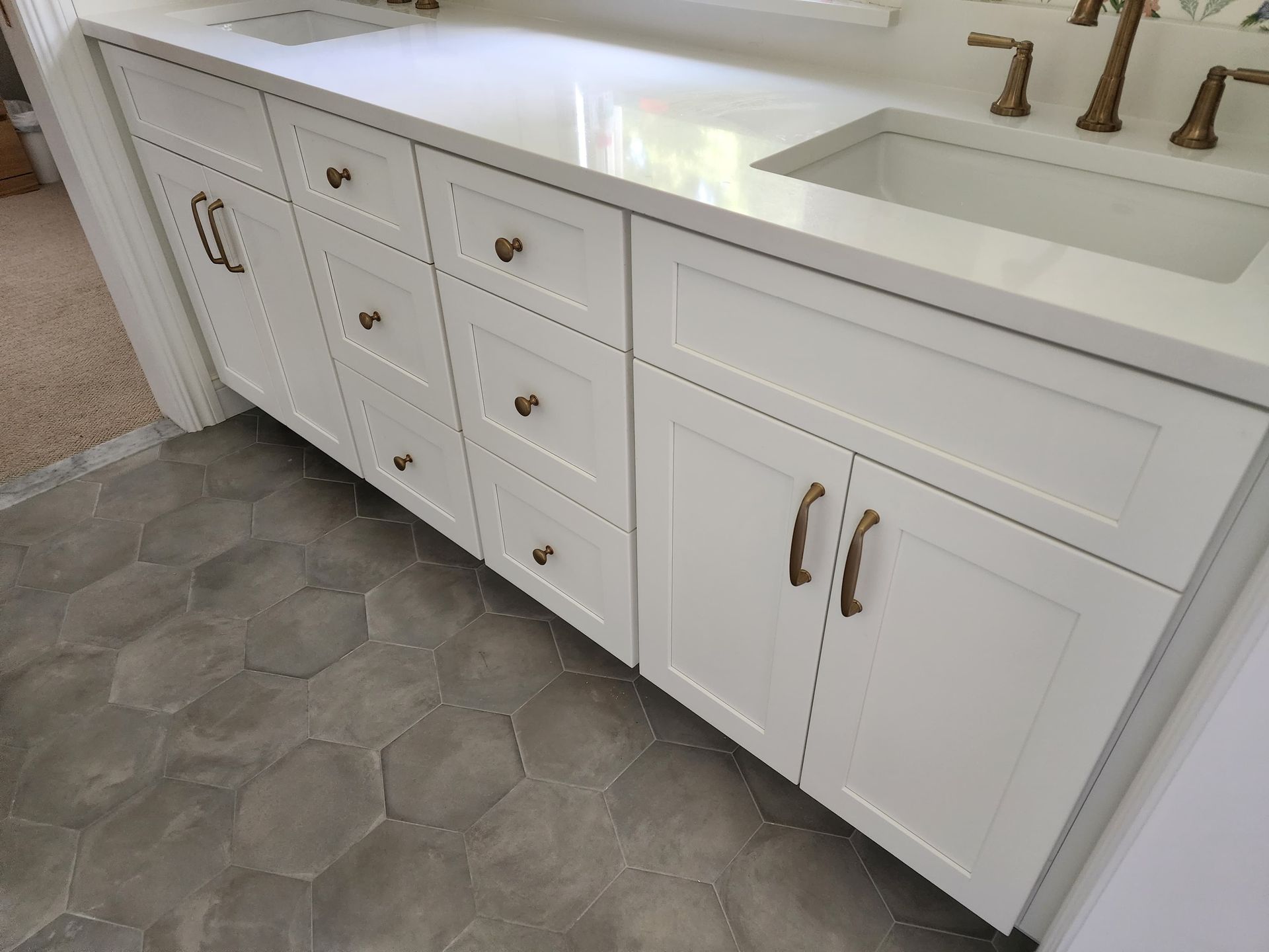 White bathroom vanity with dual sinks, gold hardware, and shaker-style cabinets, set against grey hexagonal floor tiles.