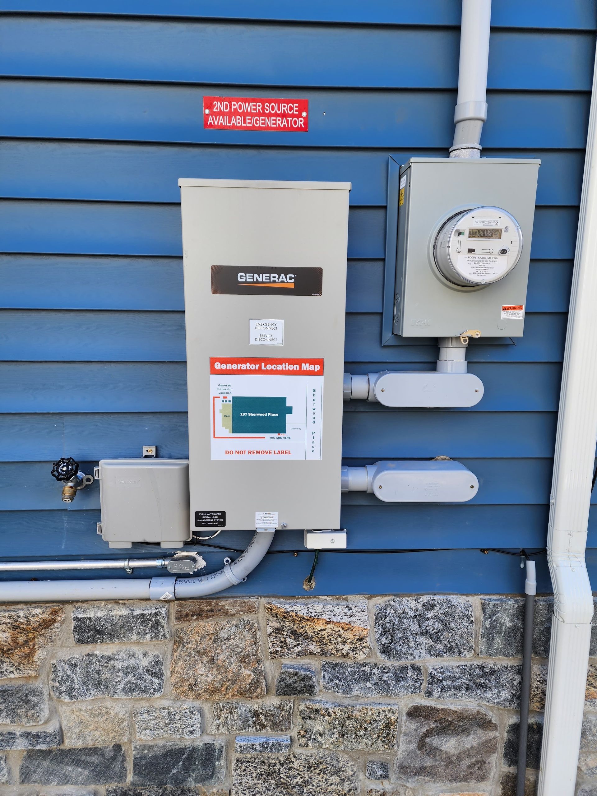 An electrical meter and a solar power inverter box mounted on the side of a blue-sided building above a stone foundation.