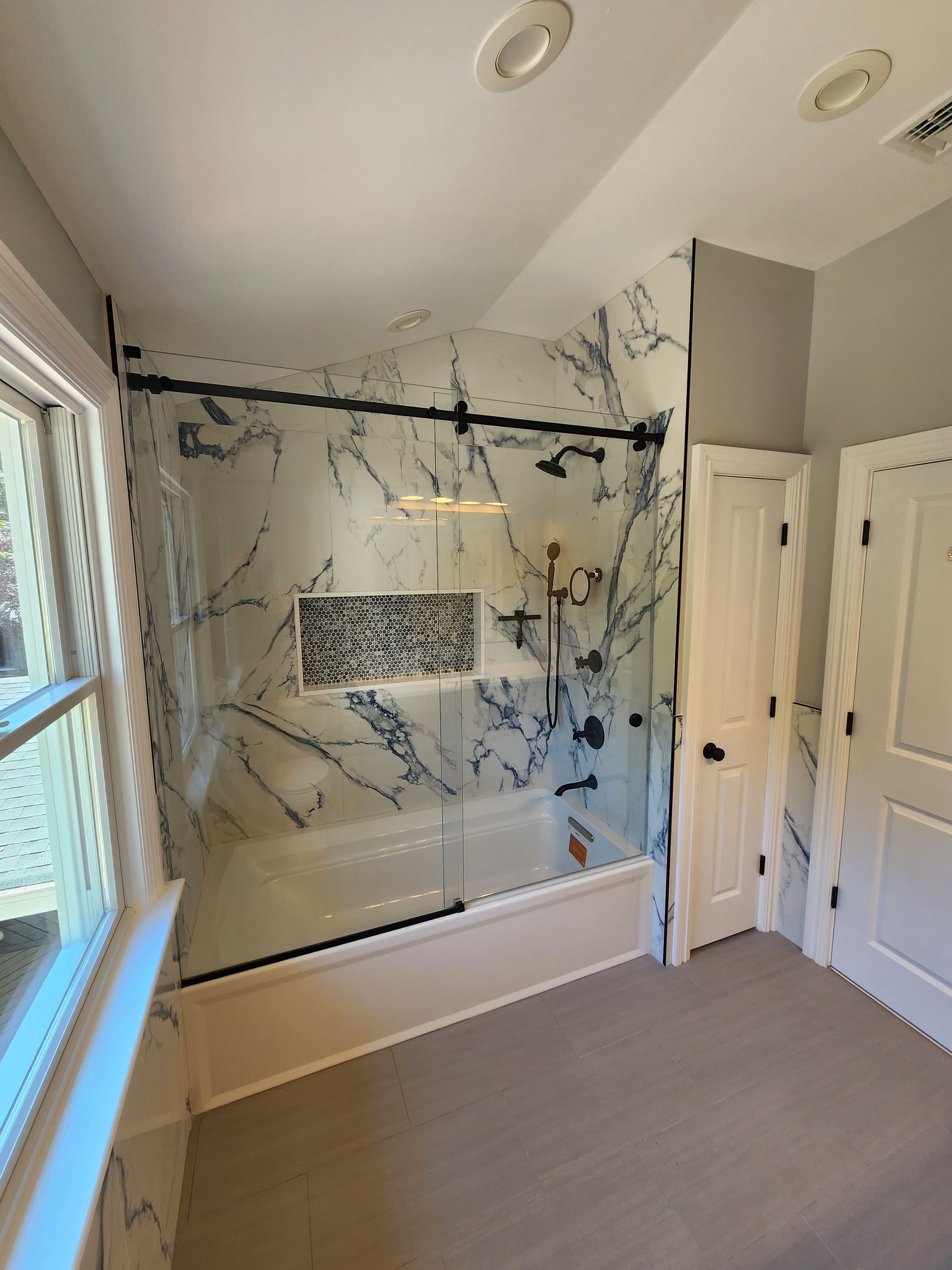 A renovated bathroom featuring a bathtub with a marble-patterned tiled surround, glass sliding doors, and black fixtures.
