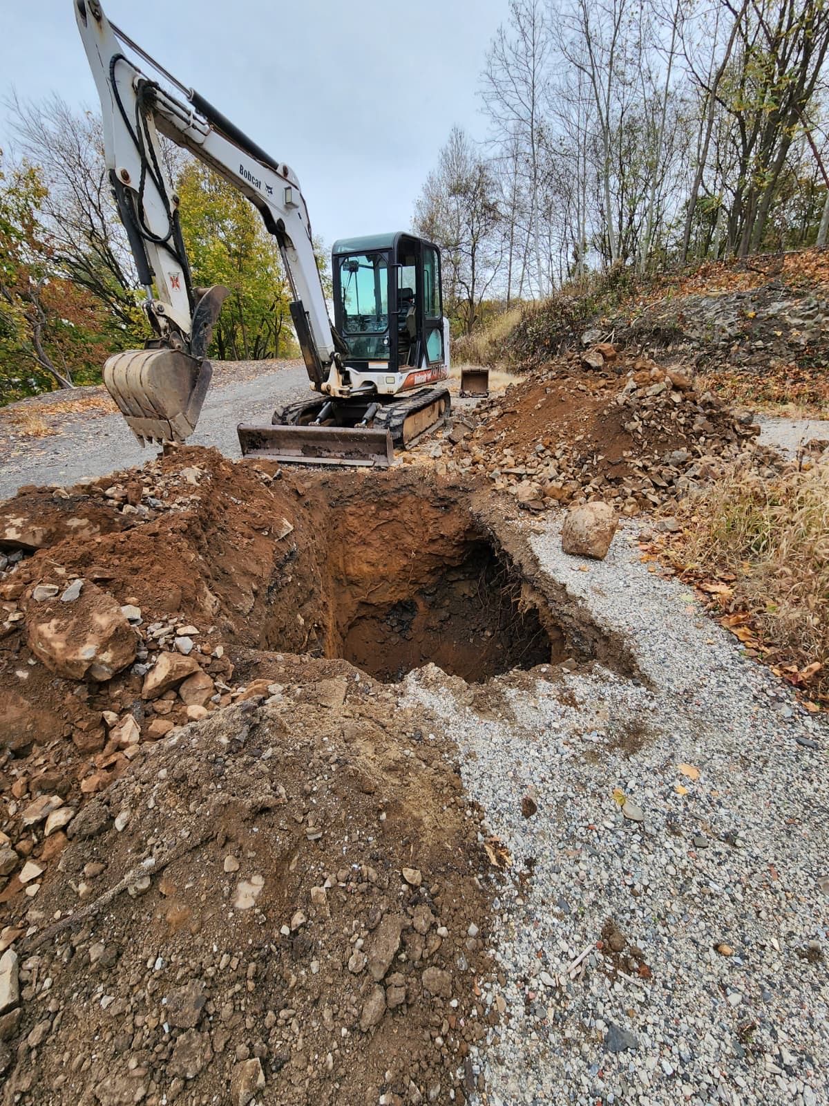 A white compact excavator sits parked on a gravel path next to a freshly dug, rectangular hole in a rocky, wooded area.
