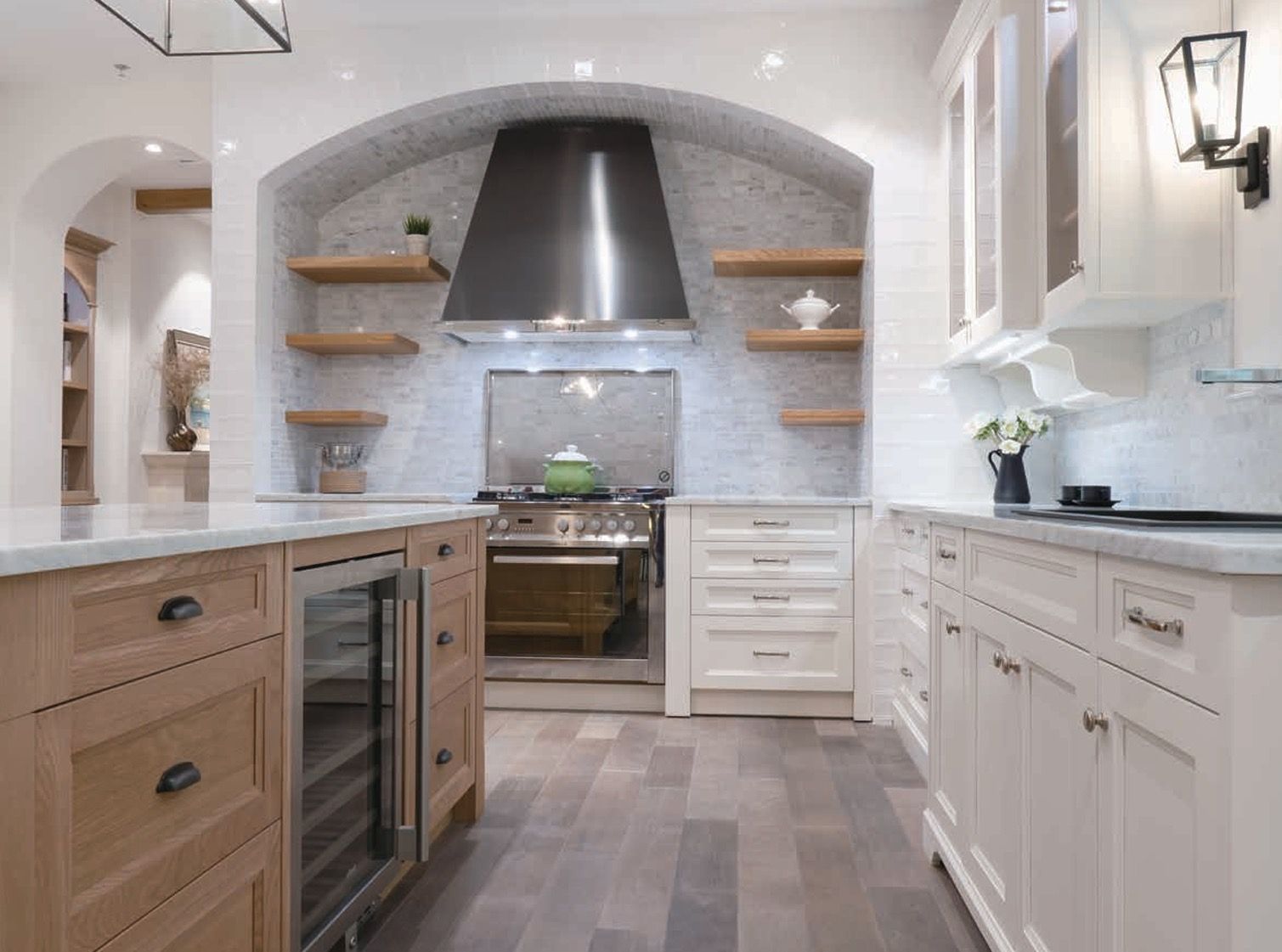 A bright kitchen featuring white cabinets, a large stainless steel range hood, wooden floating shelves, and a center island.