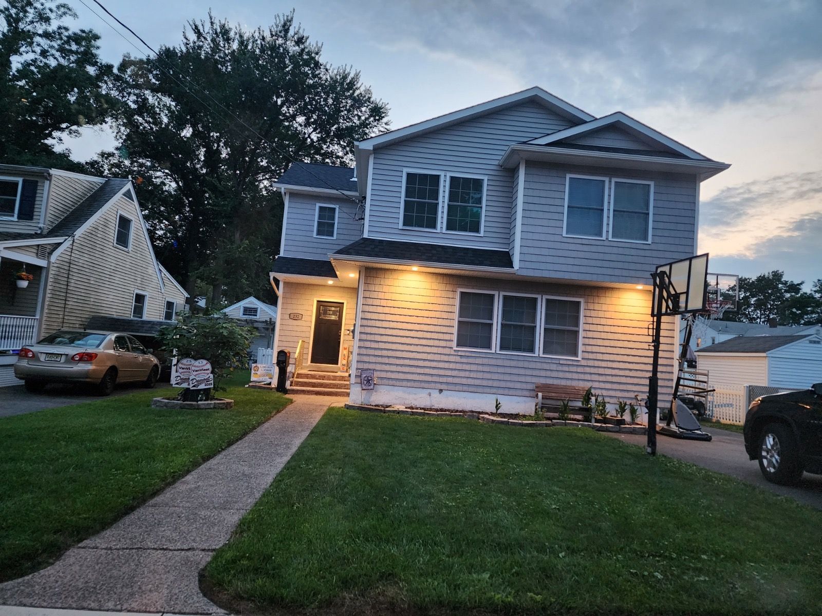 A two-story light blue house with a lit front entrance, a front lawn, and a sidewalk at dusk.
