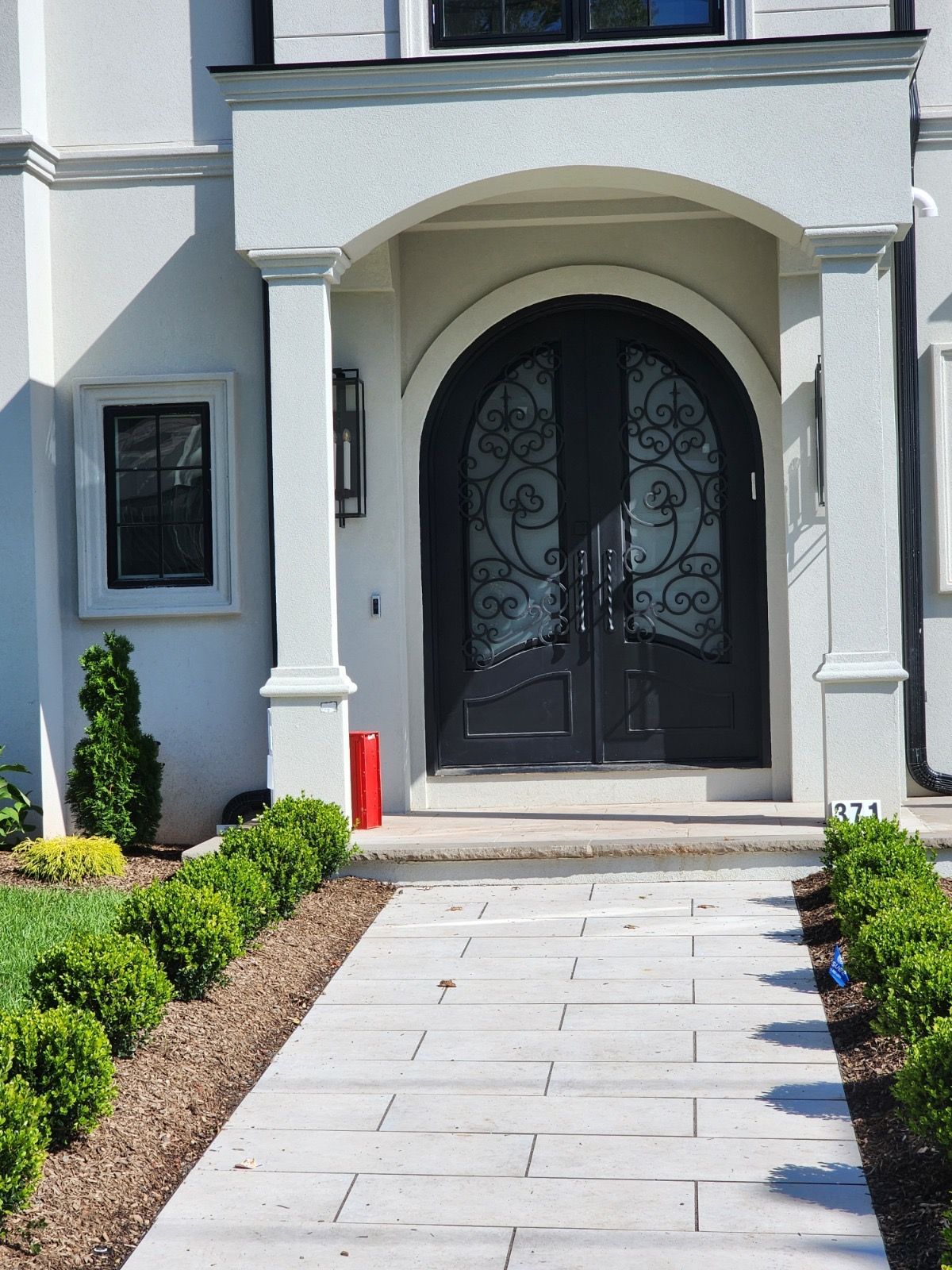 A paved walkway leads to the arched entryway of a white home with decorative black double doors and symmetrical shrubs.