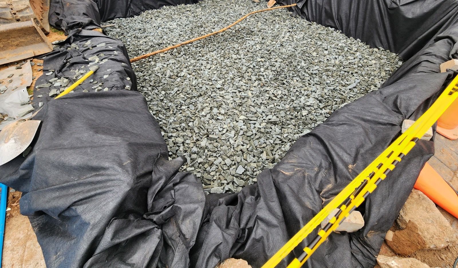 An excavation pit lined with black landscape fabric and partially filled with grey gravel, surrounded by yellow tape.