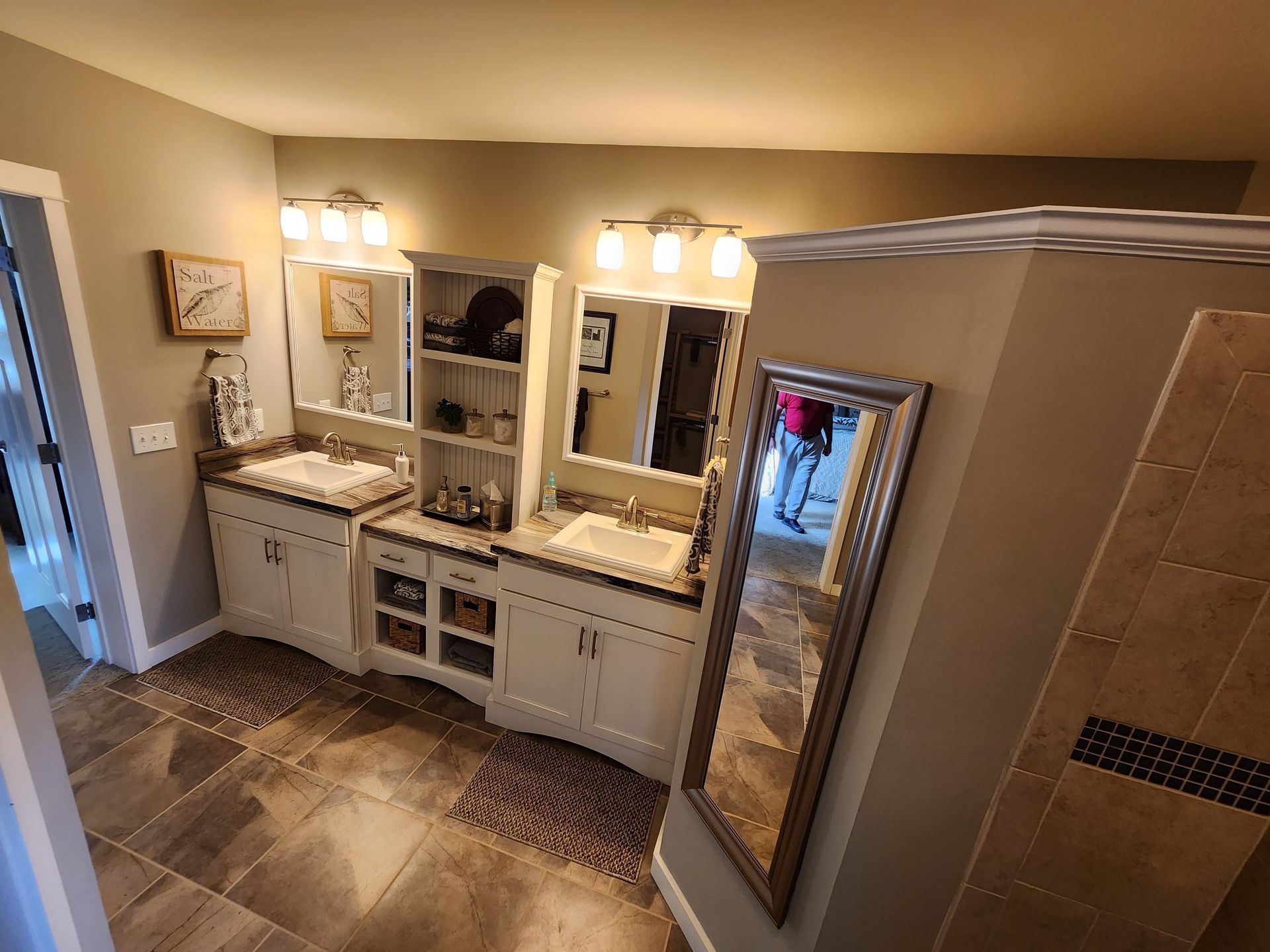 A double vanity bathroom with white cabinets, beige walls, tiled floors, a tower shelf, and a wall-mounted mirror.