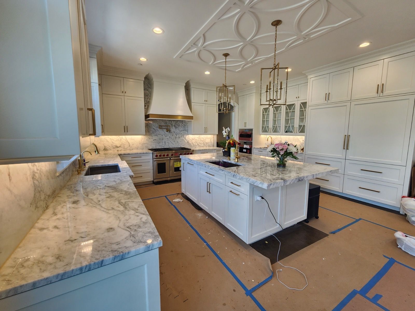 A bright kitchen with white cabinets, a large marble-topped island, gold fixtures, and protective floor covering.