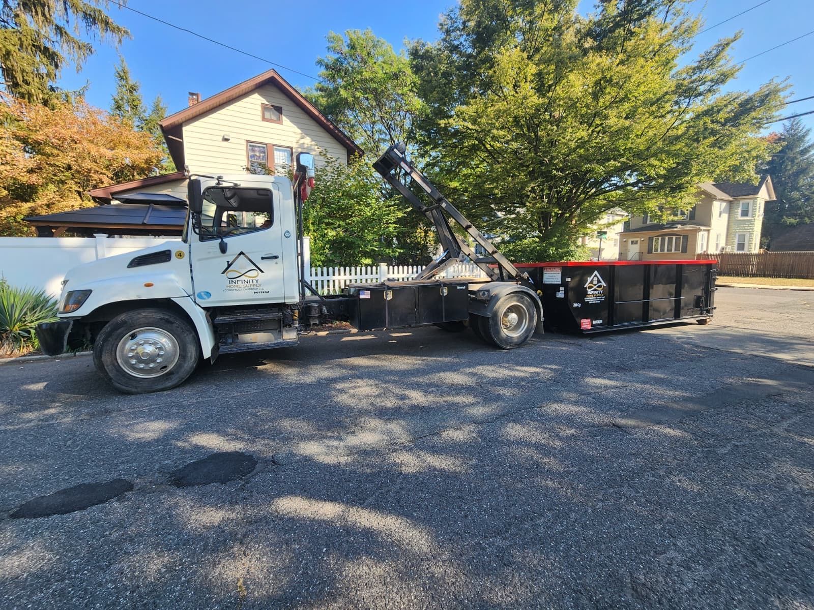 A white roll-off dump truck with a black container parked on a gravel lot in front of houses and trees.