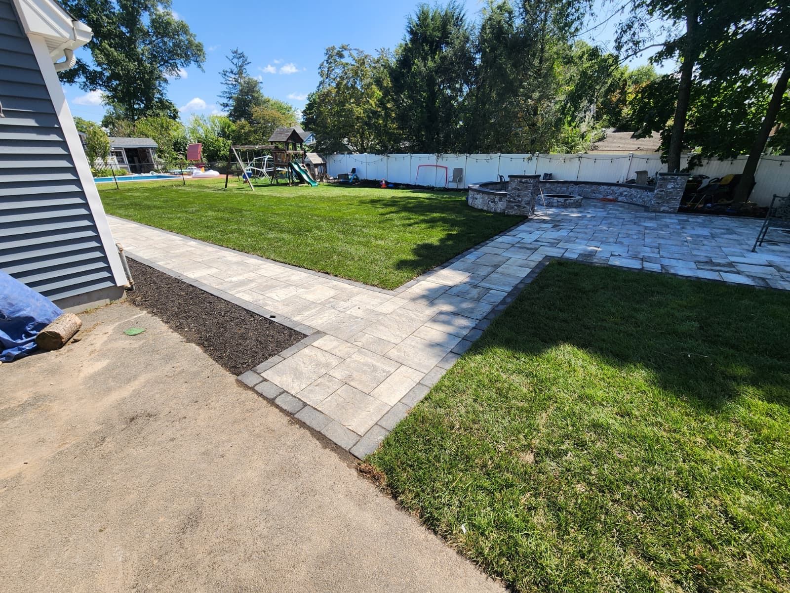 A backyard featuring a stone paver path connecting a concrete patio to a larger stone area with a retaining wall.