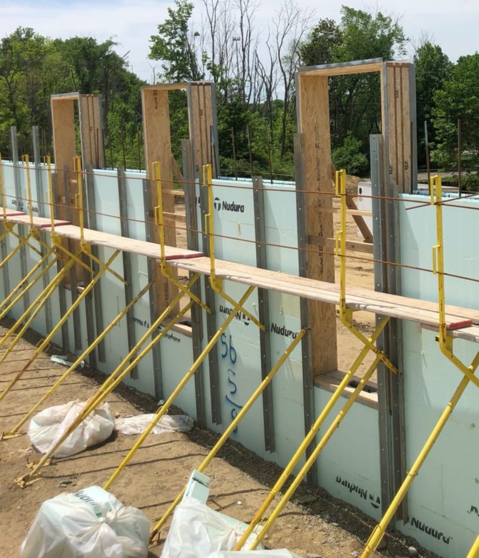 Construction site showing an insulated concrete form wall with wooden window frames and yellow scaffolding brackets.