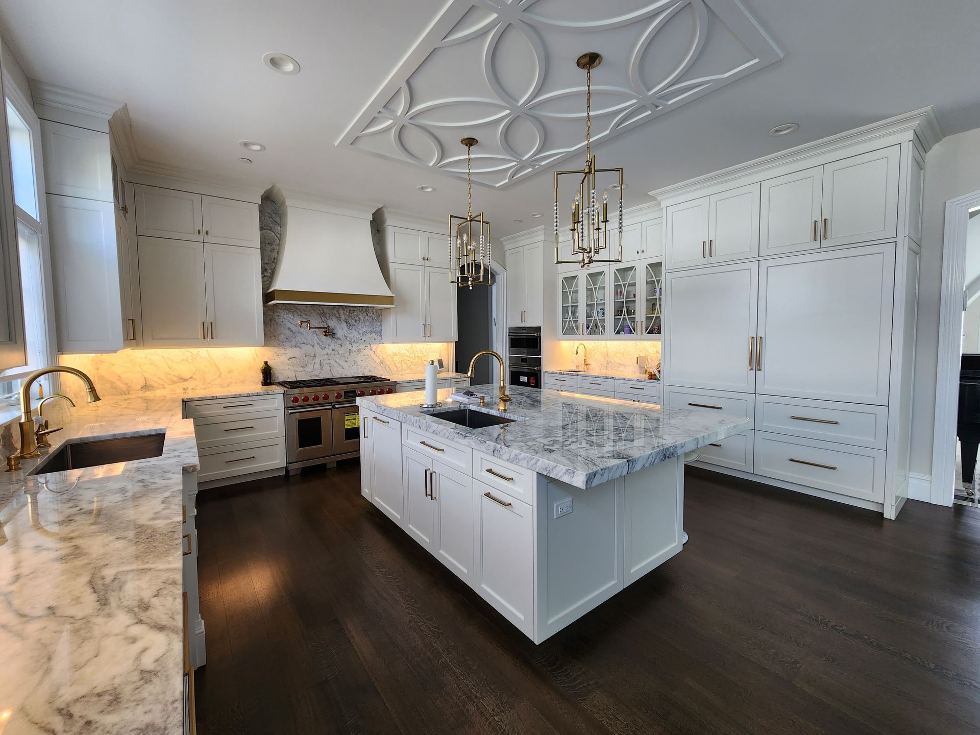 Modern, white kitchen with marble countertops, a central island, gold light fixtures, and dark hardwood floors.