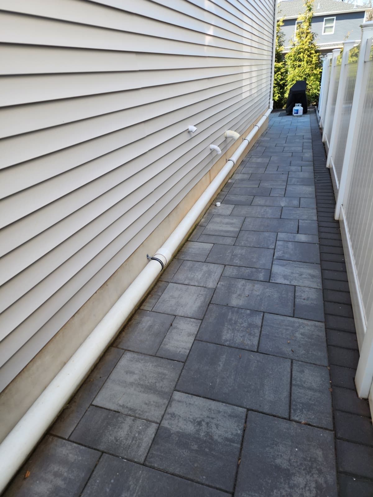 A narrow outdoor walkway paved with gray stone bricks, flanked by beige vinyl siding on one side and a white fence.