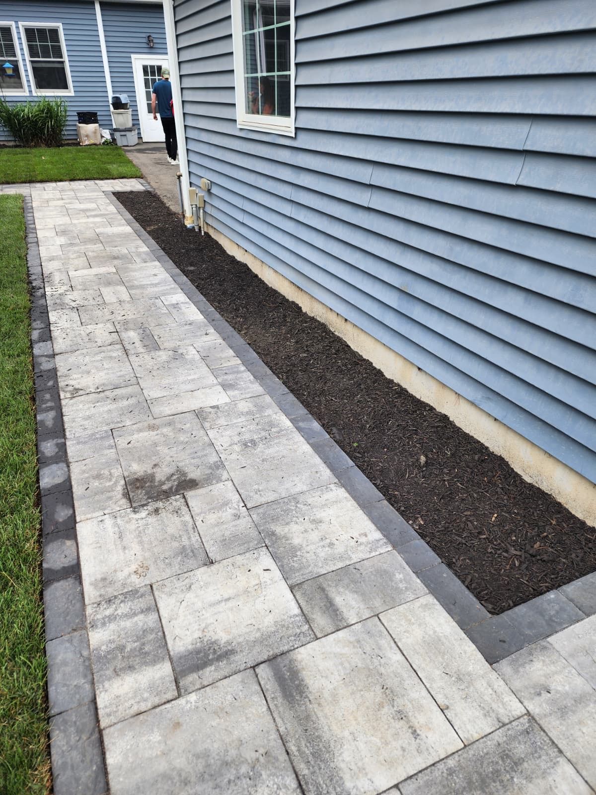 A grey paver walkway runs alongside a blue-sided house, separated by a narrow strip of dark mulch.