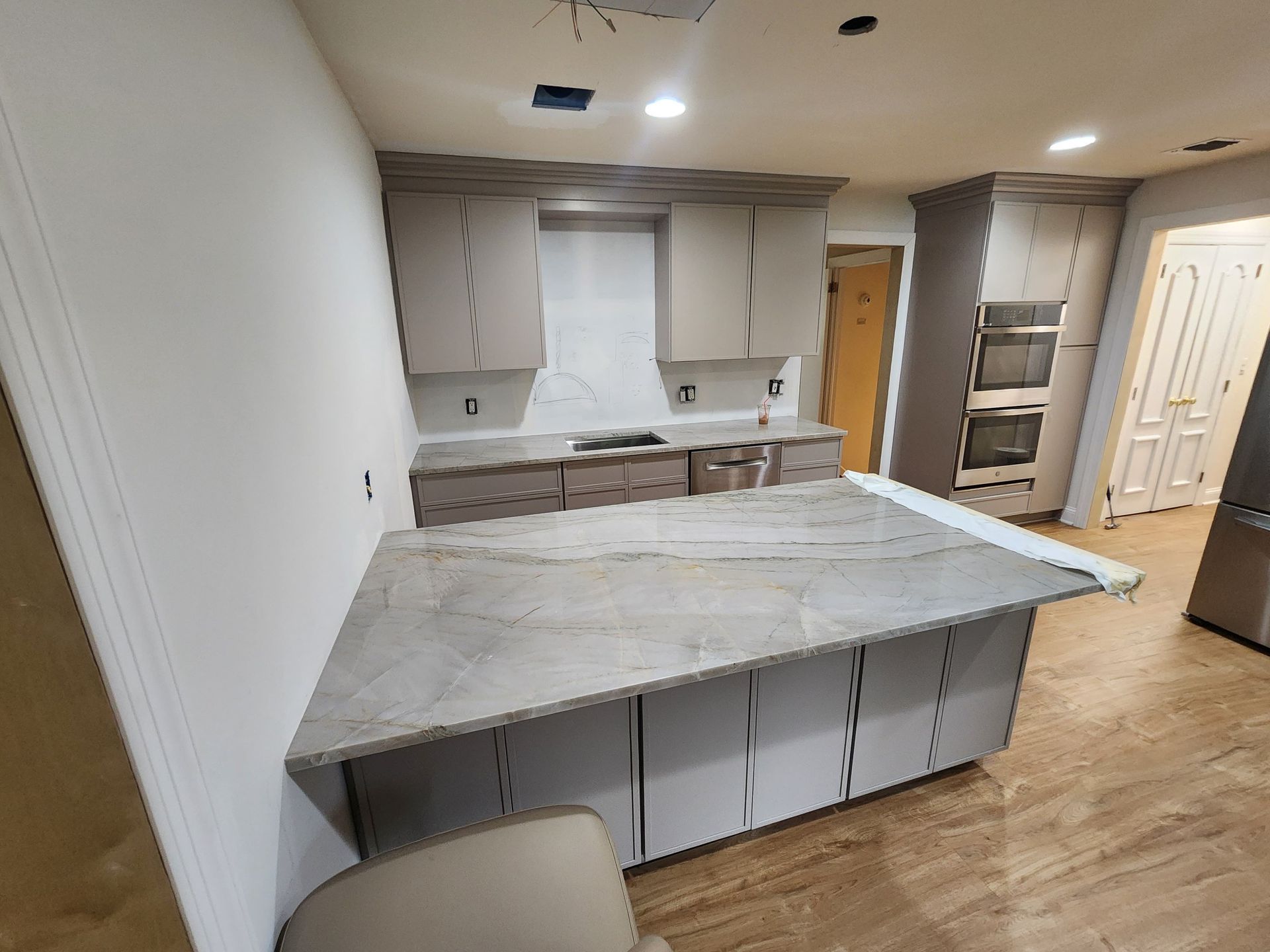 A kitchen featuring a large stone-topped island, light-colored cabinets, and built-in double ovens on a wood floor.