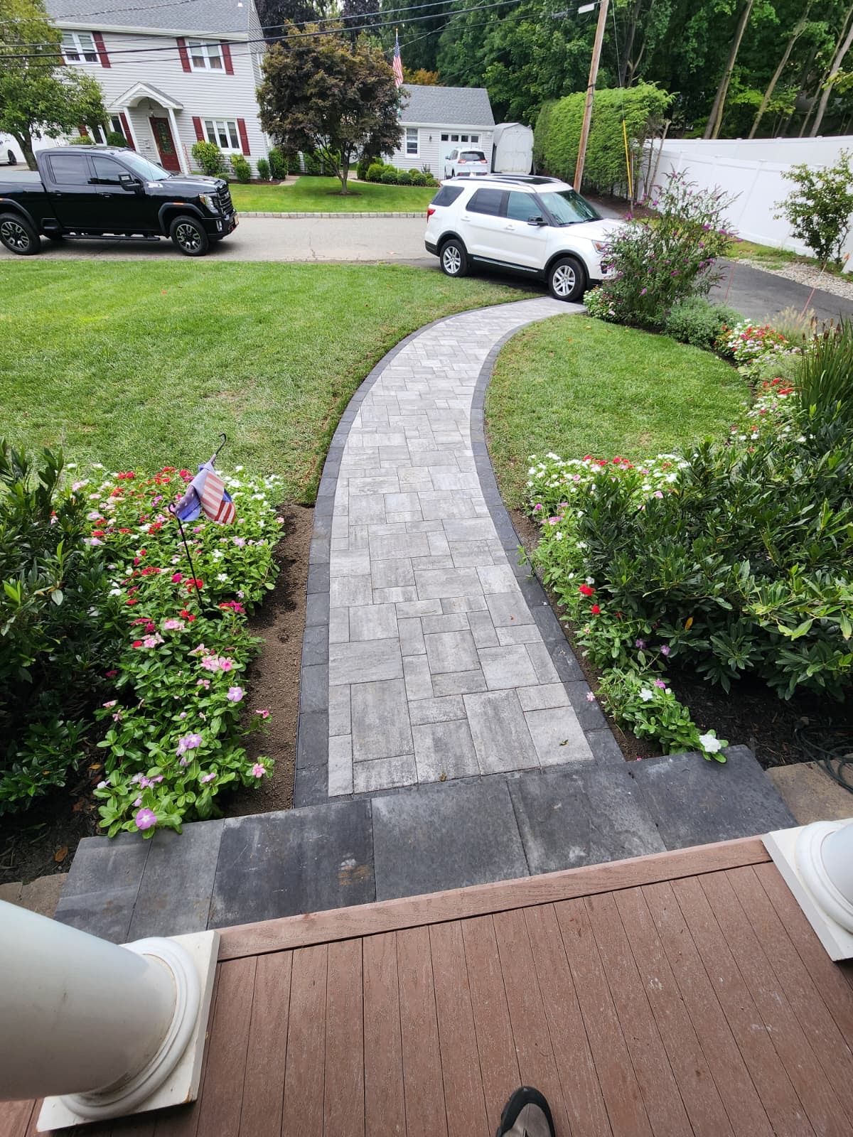 A curved, gray paver walkway leads from a wooden porch across a lawn toward a driveway with two parked vehicles.