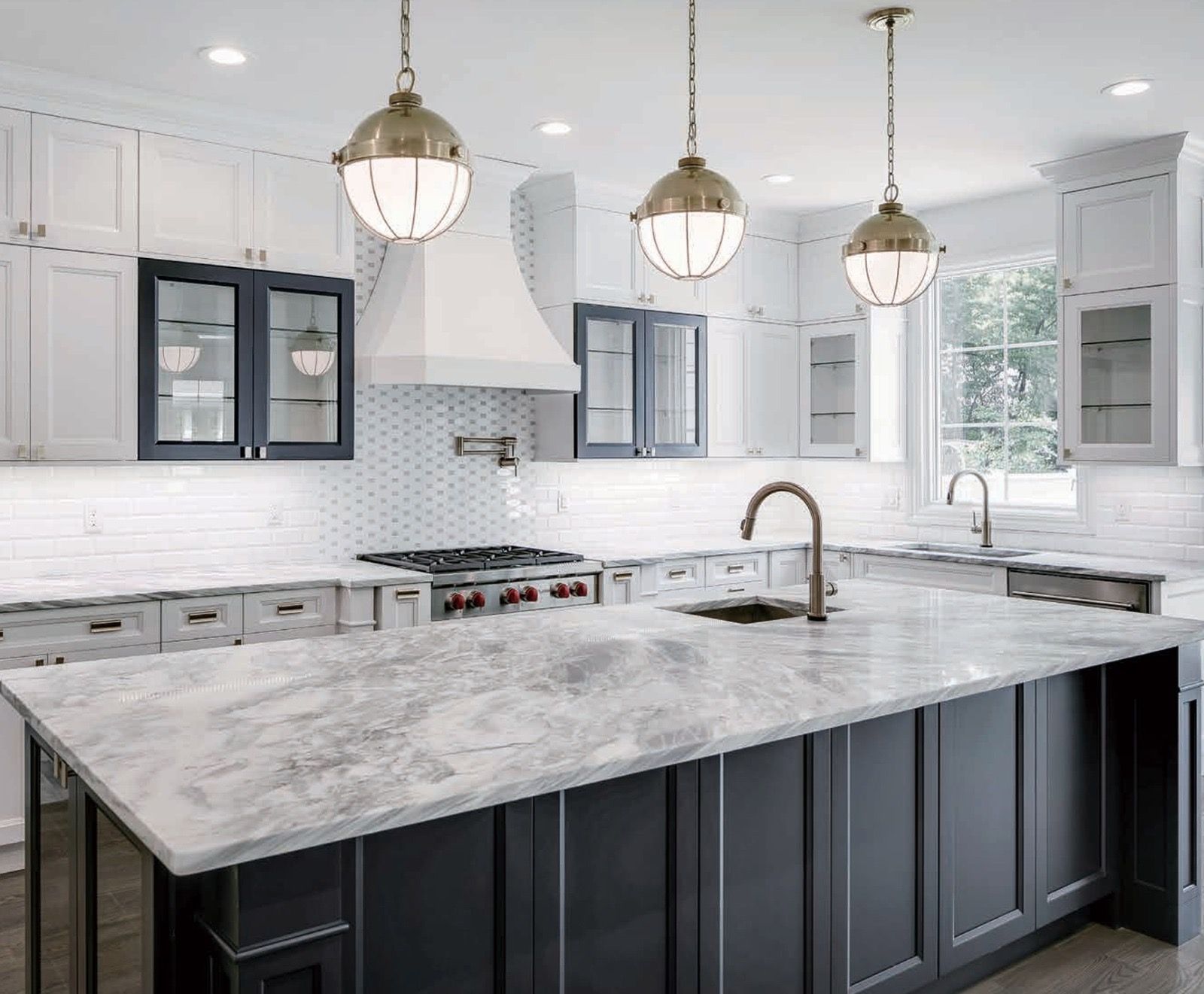 A bright kitchen with white and dark blue cabinetry, a large marble-topped island, and three hanging brass pendant lights.