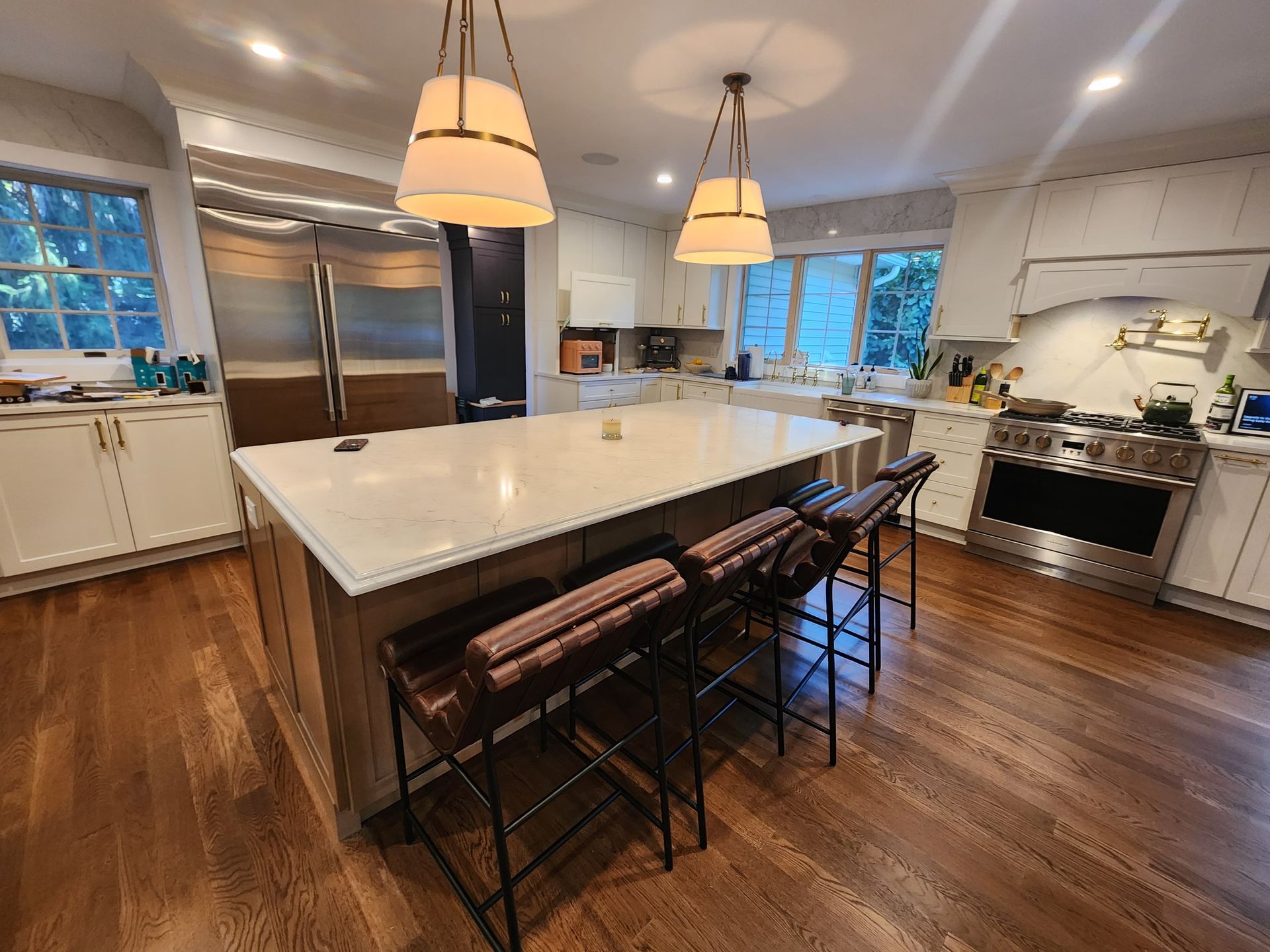 A modern, well-lit kitchen with a large central island, three bar stools, white cabinetry, and stainless steel appliances.