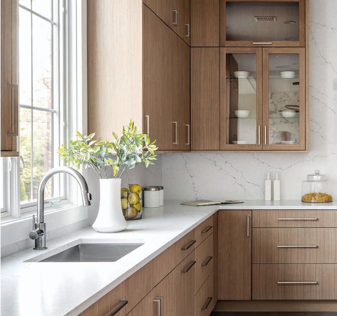 A kitchen with light wood cabinets, white countertops, a stainless steel sink, and a glass-front upper cabinet.