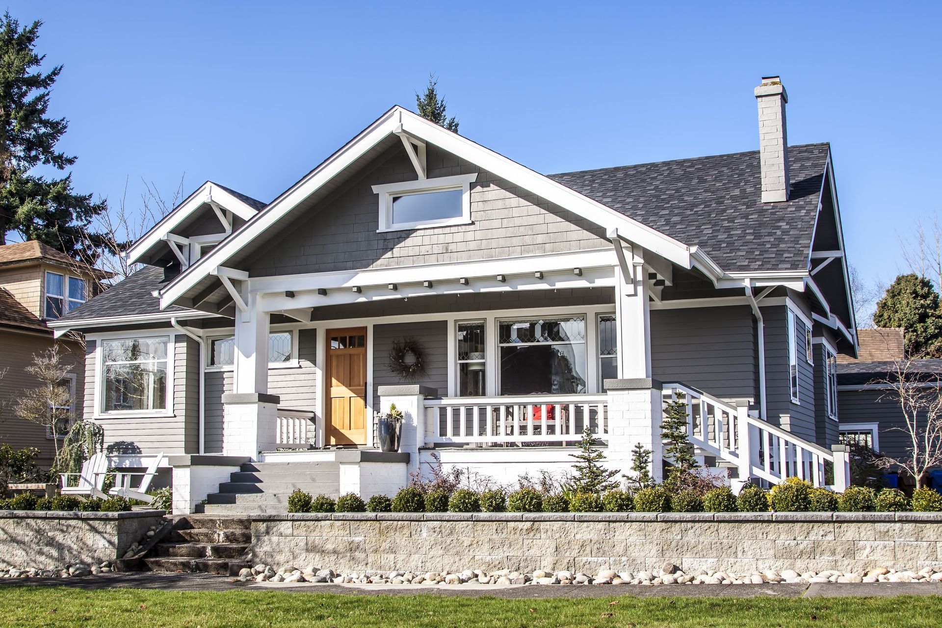 A gray-sided craftsman-style house with a covered front porch, white trim, and stone retaining wall under a blue sky.