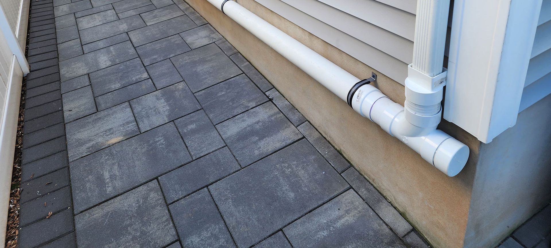 A white downspout extension connected to a PVC pipe running horizontally along a house foundation next to stone pavers.