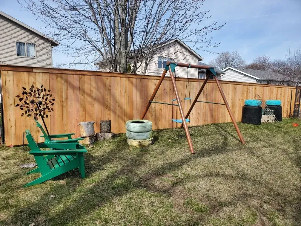 A wooden fence with a swing set and a green chair in the backyard.
