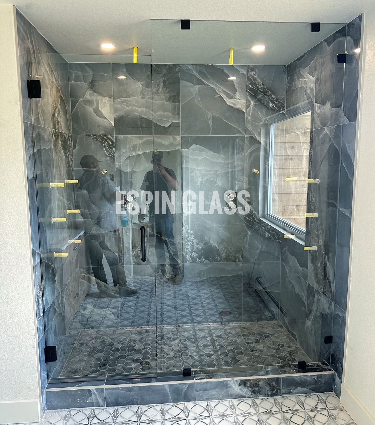 Glass shower enclosure with dark marble tiles. Two people reflected in the glass.