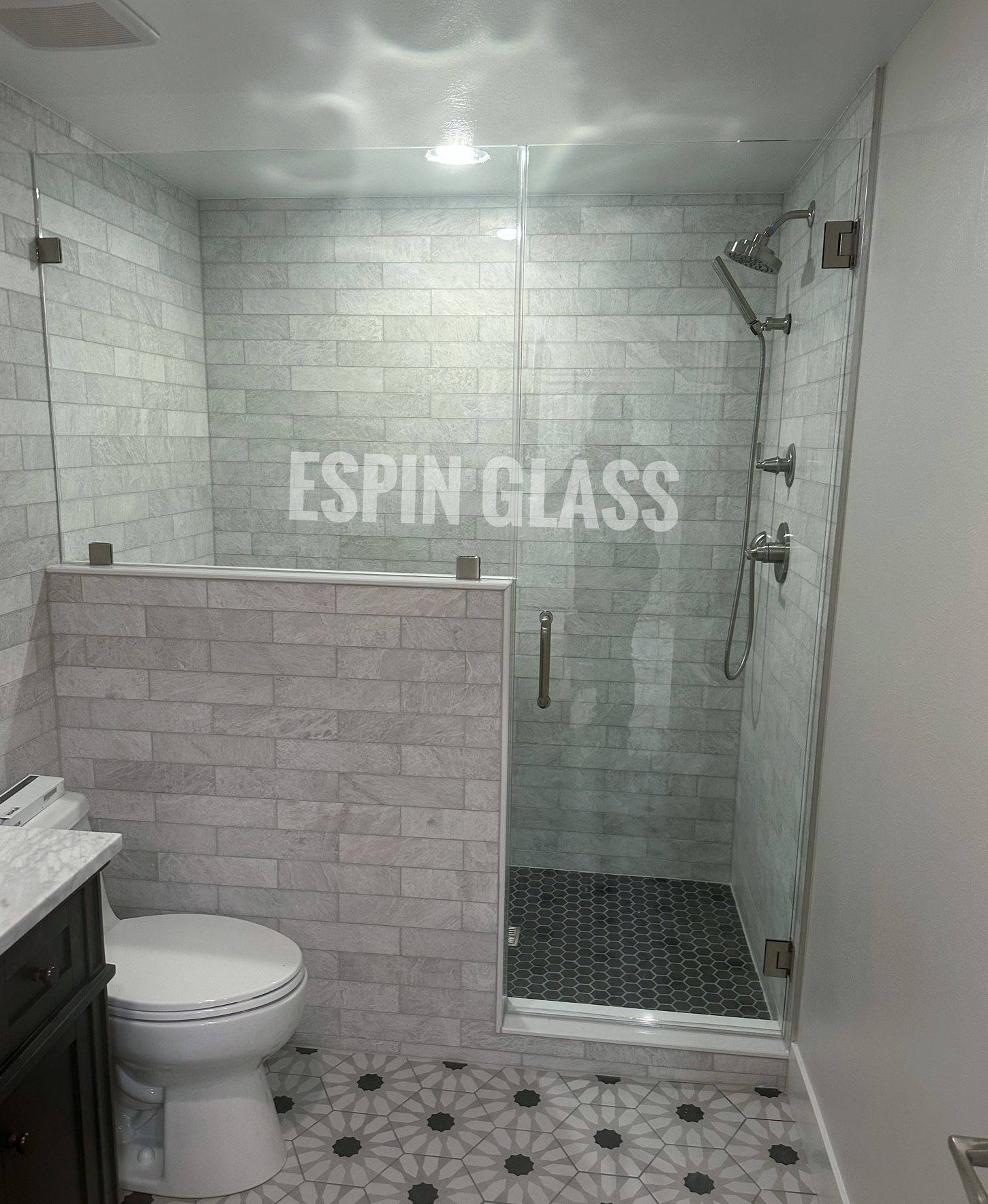 Glass shower enclosure with gray stone tile walls, black tiled shower floor, and patterned floor.