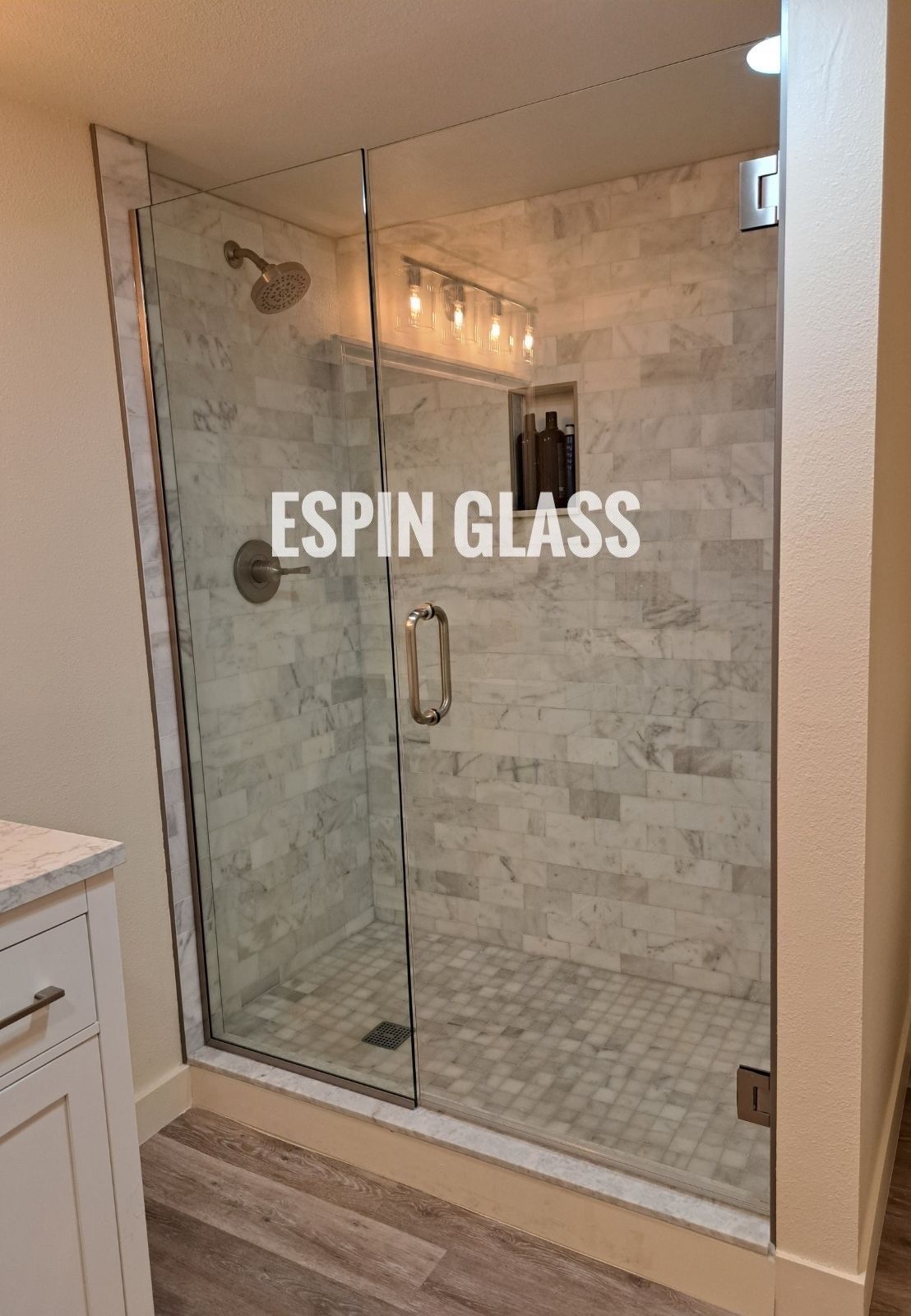 Frameless glass shower enclosure with white marble tiles, silver fixtures, and gray and white flooring.