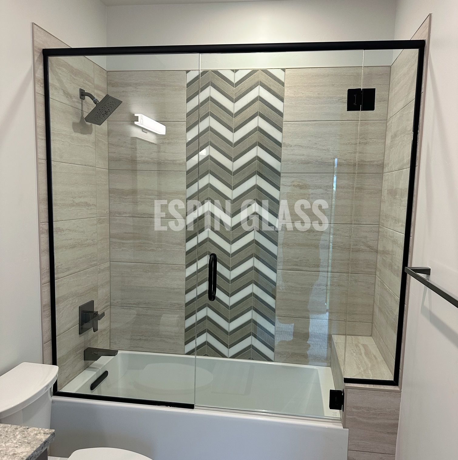 Modern bathroom with a black-framed glass shower enclosure over a white bathtub, featuring patterned gray tiles.