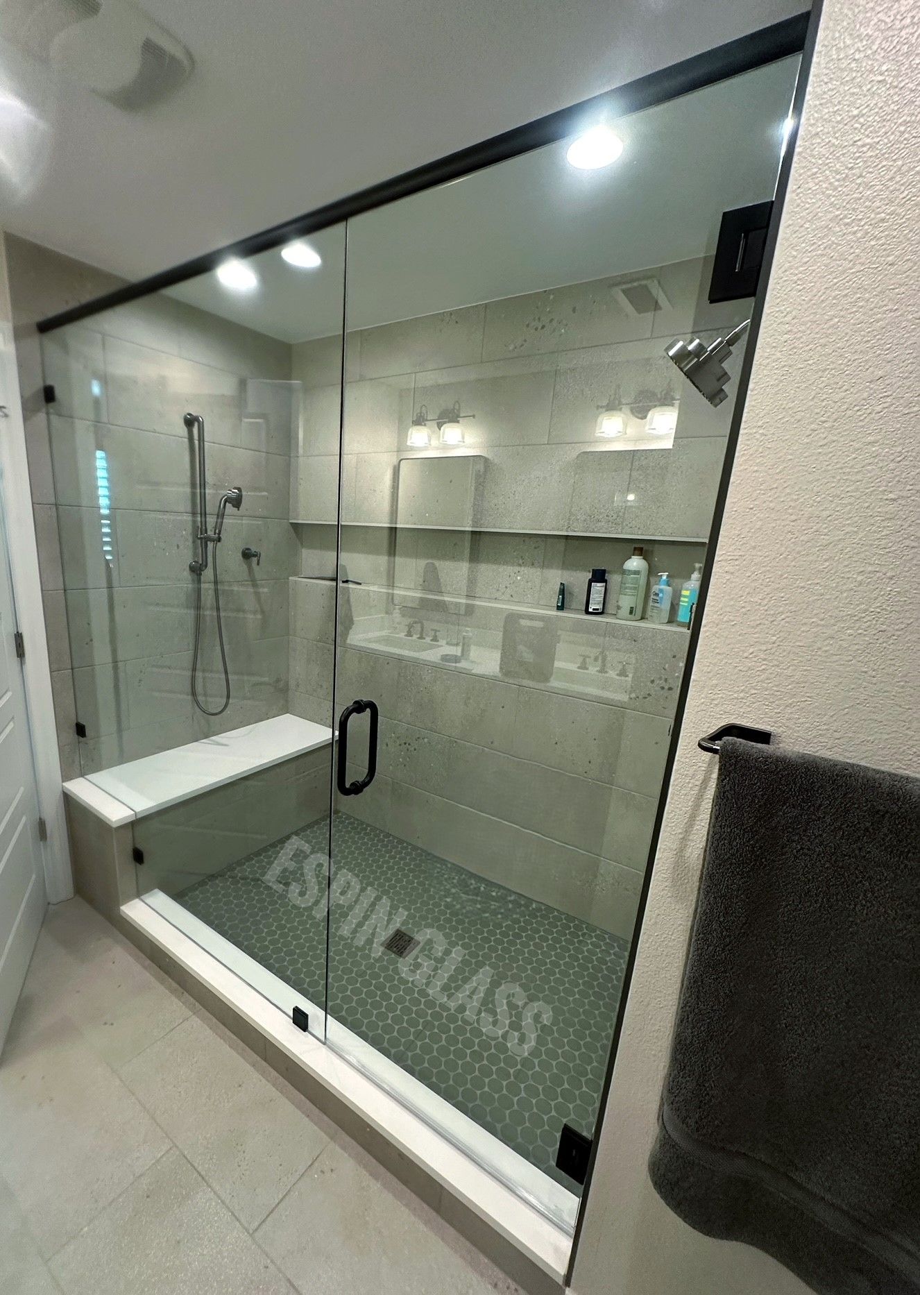 Modern walk-in shower with glass panels, black hardware, and built-in bench. Gray tiles, showerhead, and towels.