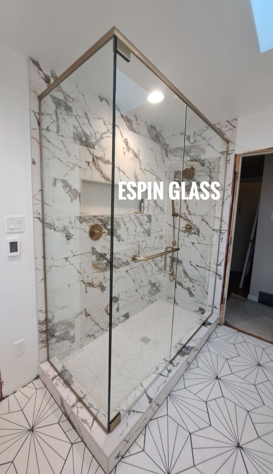 A glass shower enclosure with gold trim in a marble-tiled bathroom; ESPIN GLASS branding is visible.