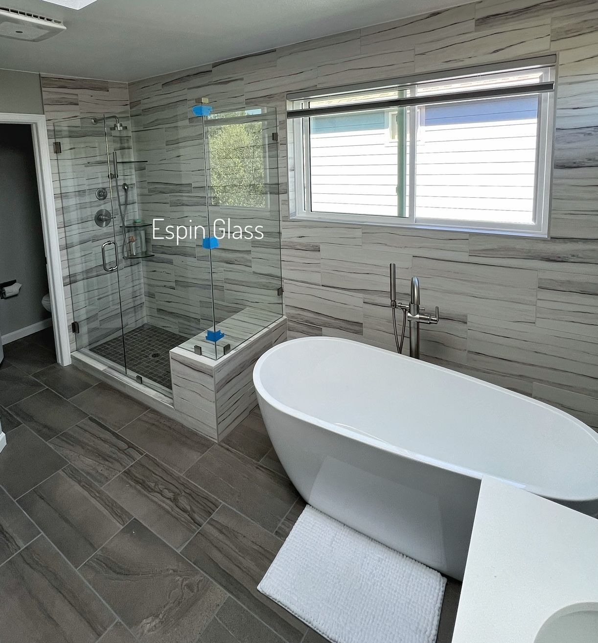 Modern bathroom with glass shower, white tub, wood-look tile, and a window with blinds.