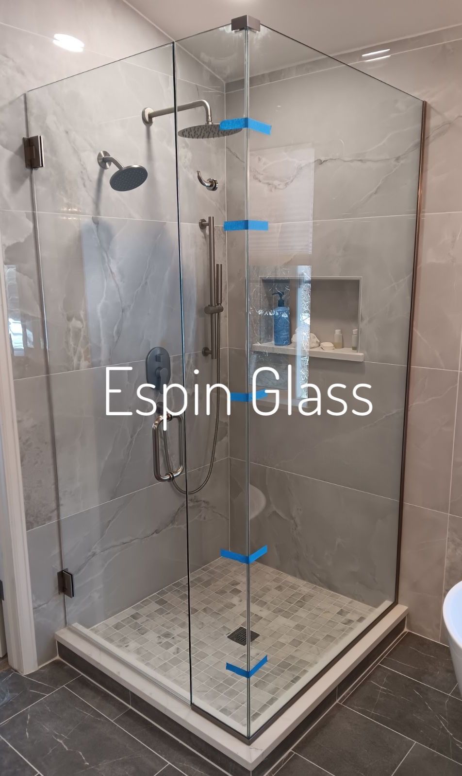 Frameless glass shower enclosure with gray tile walls and floor; showerheads, and a niche with toiletries.