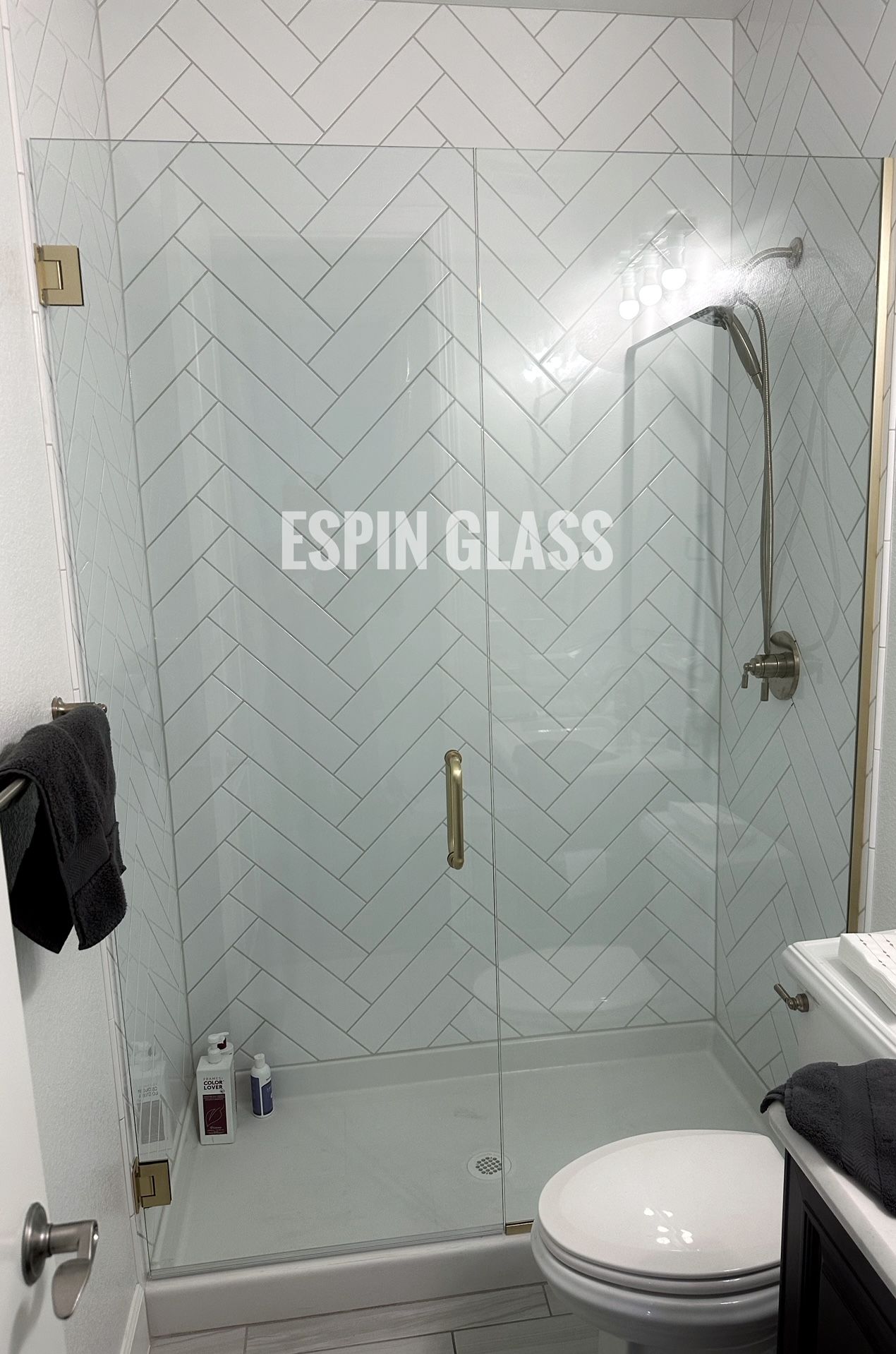 Shower with glass door and gold hardware against a white herringbone tile backdrop; toilet and sink visible.