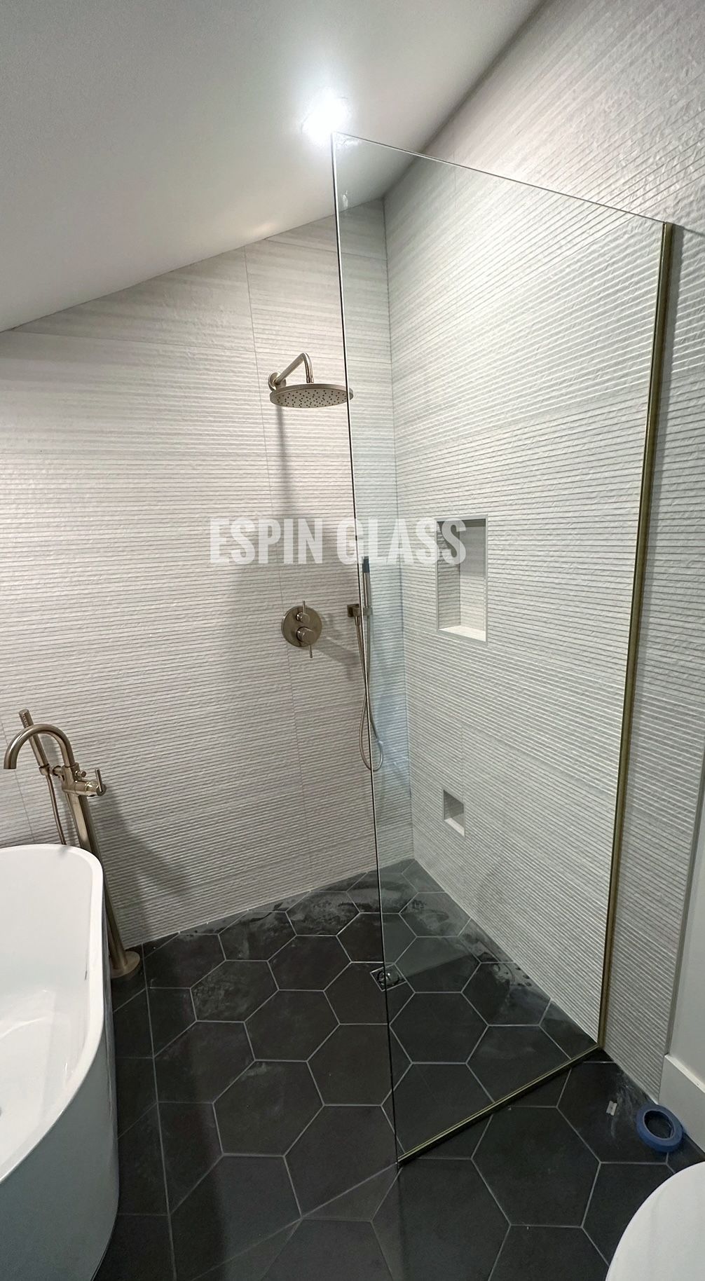 Shower with glass panel, metal fixtures, white textured wall, and dark hexagon floor tiles.