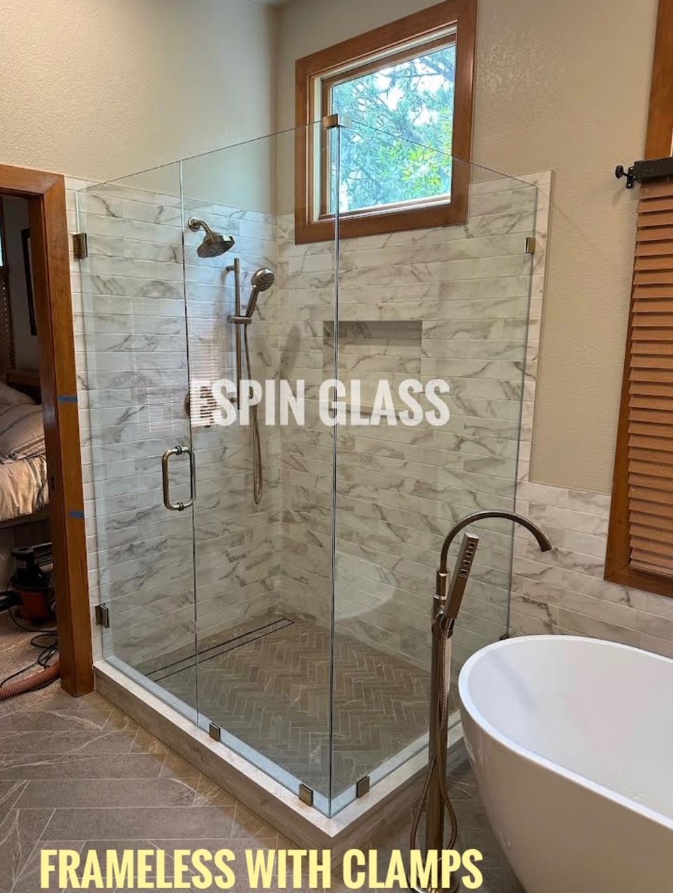 Frameless glass shower with marble tile walls and a window above. Has a bathtub and fixtures in a modern bathroom.