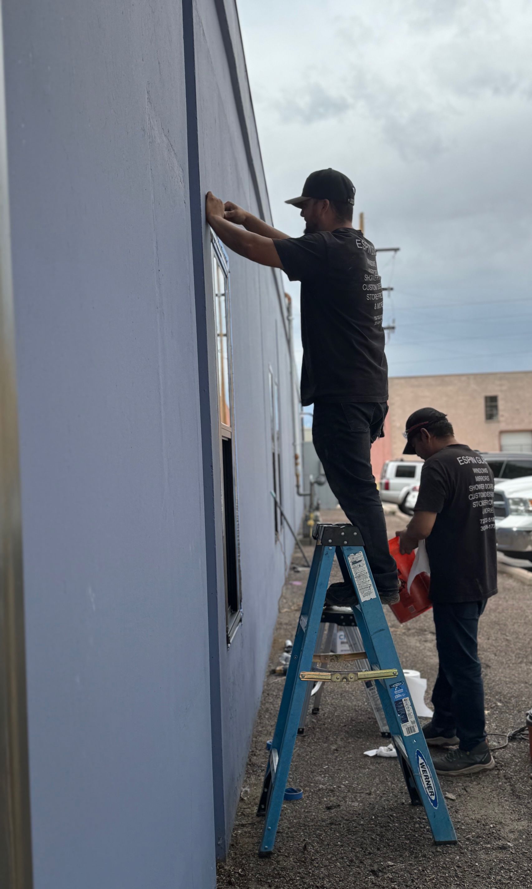 Two men painting a pale blue building. One on a ladder, the other holding a bucket. Cloudy sky.
