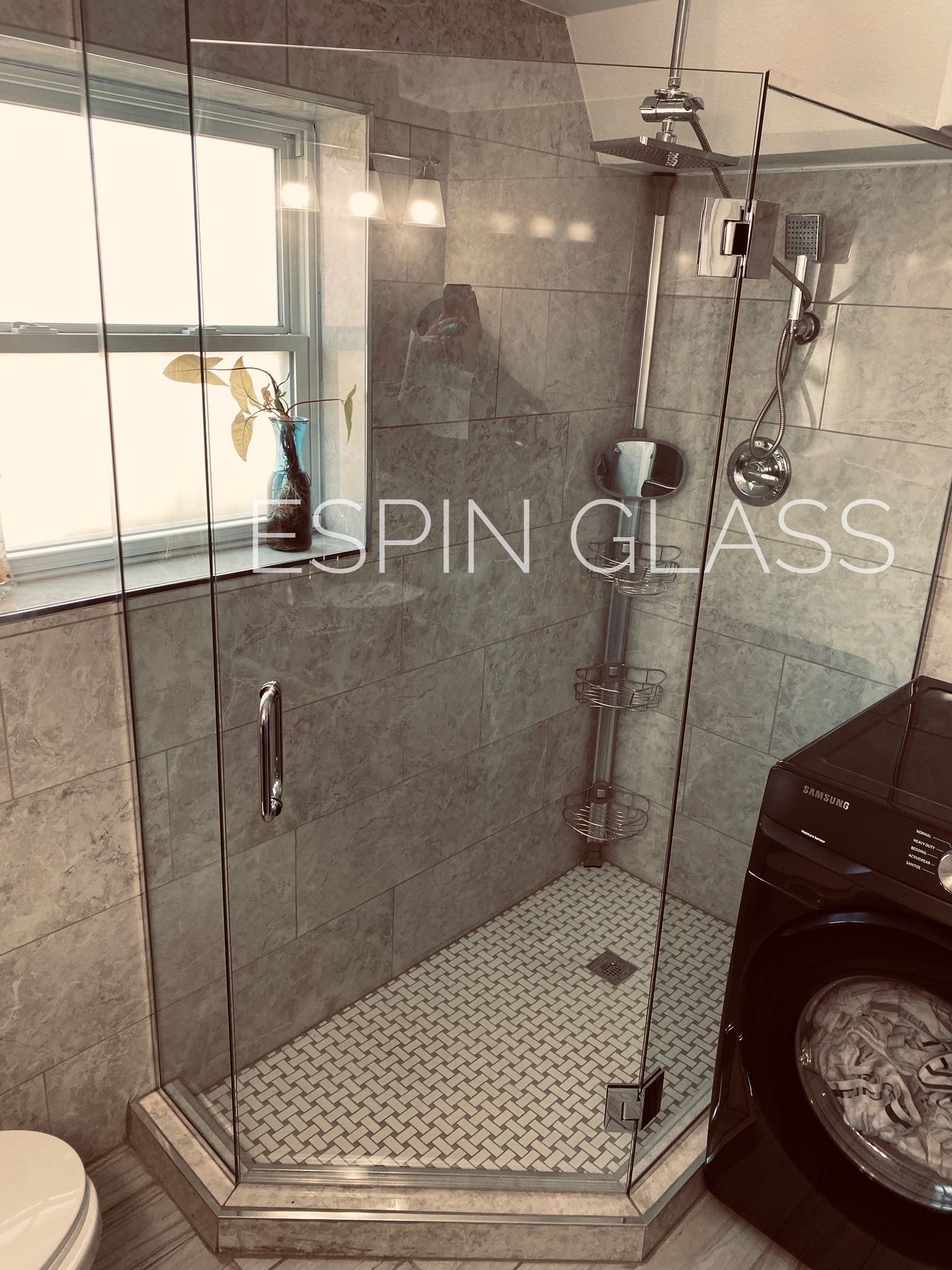 Frameless glass shower with gray tile, small white tile floor, and a dark washing machine in a bathroom.