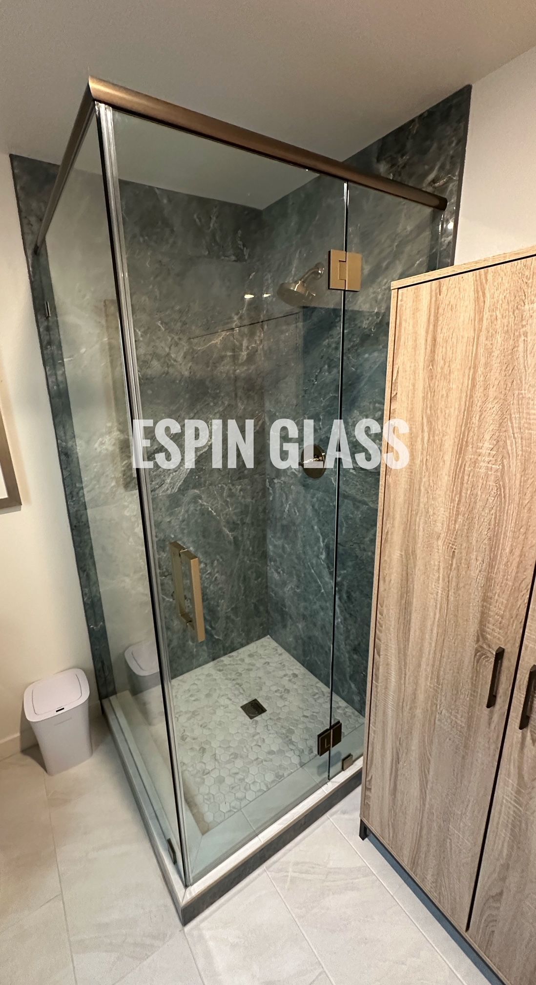 Frameless shower enclosure with bronze-colored trim and hinges, installed in a bathroom with marble-look tile.