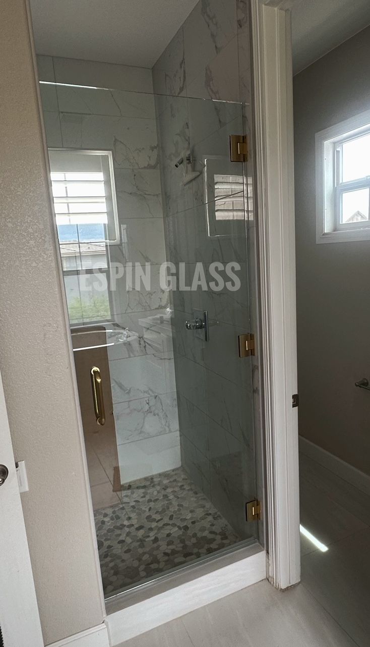 Glass shower enclosure with gold hardware, set in a light bathroom with window.
