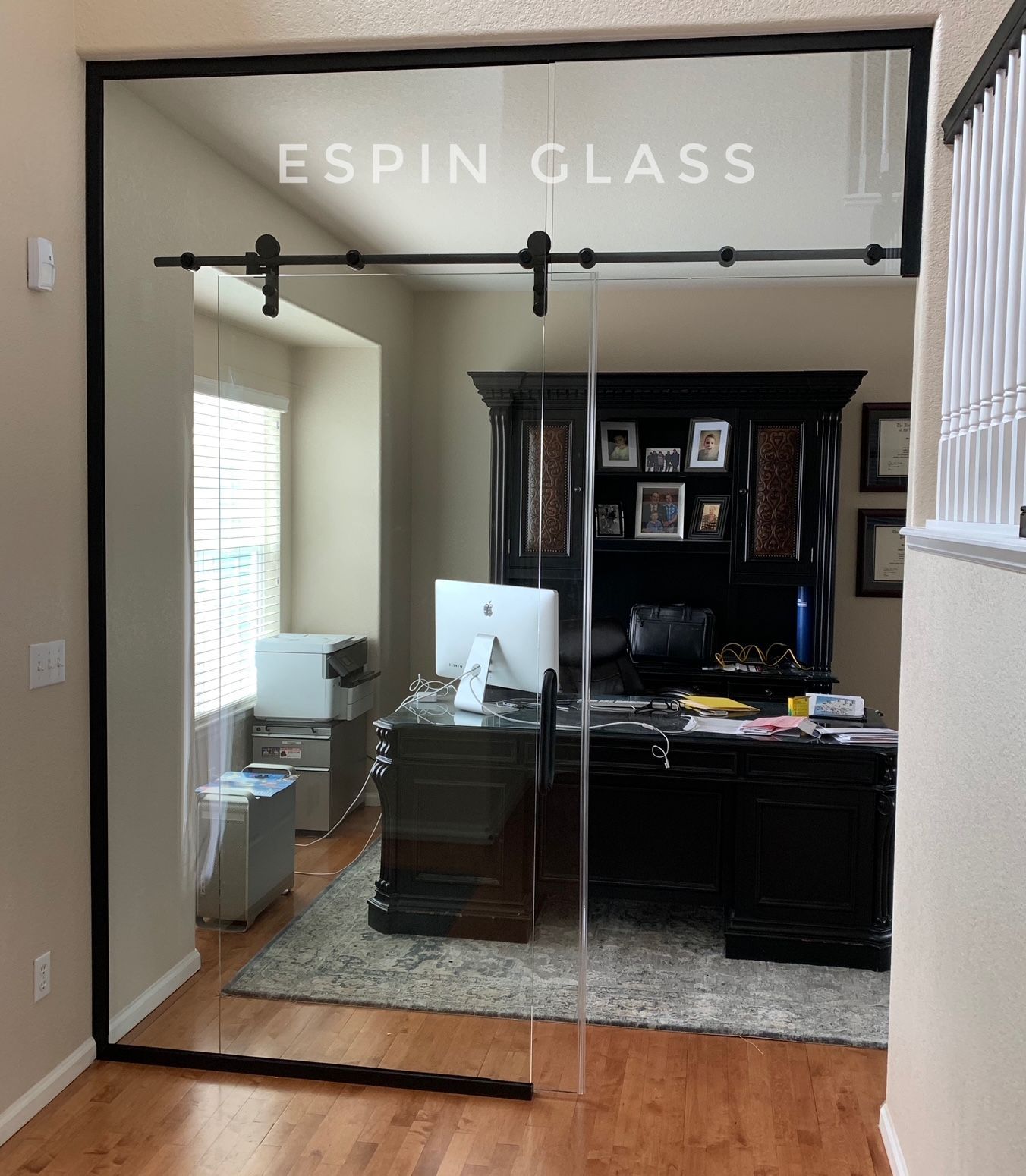 Glass sliding door separates a home office, featuring a desk and printer, from a hallway with hardwood floors.