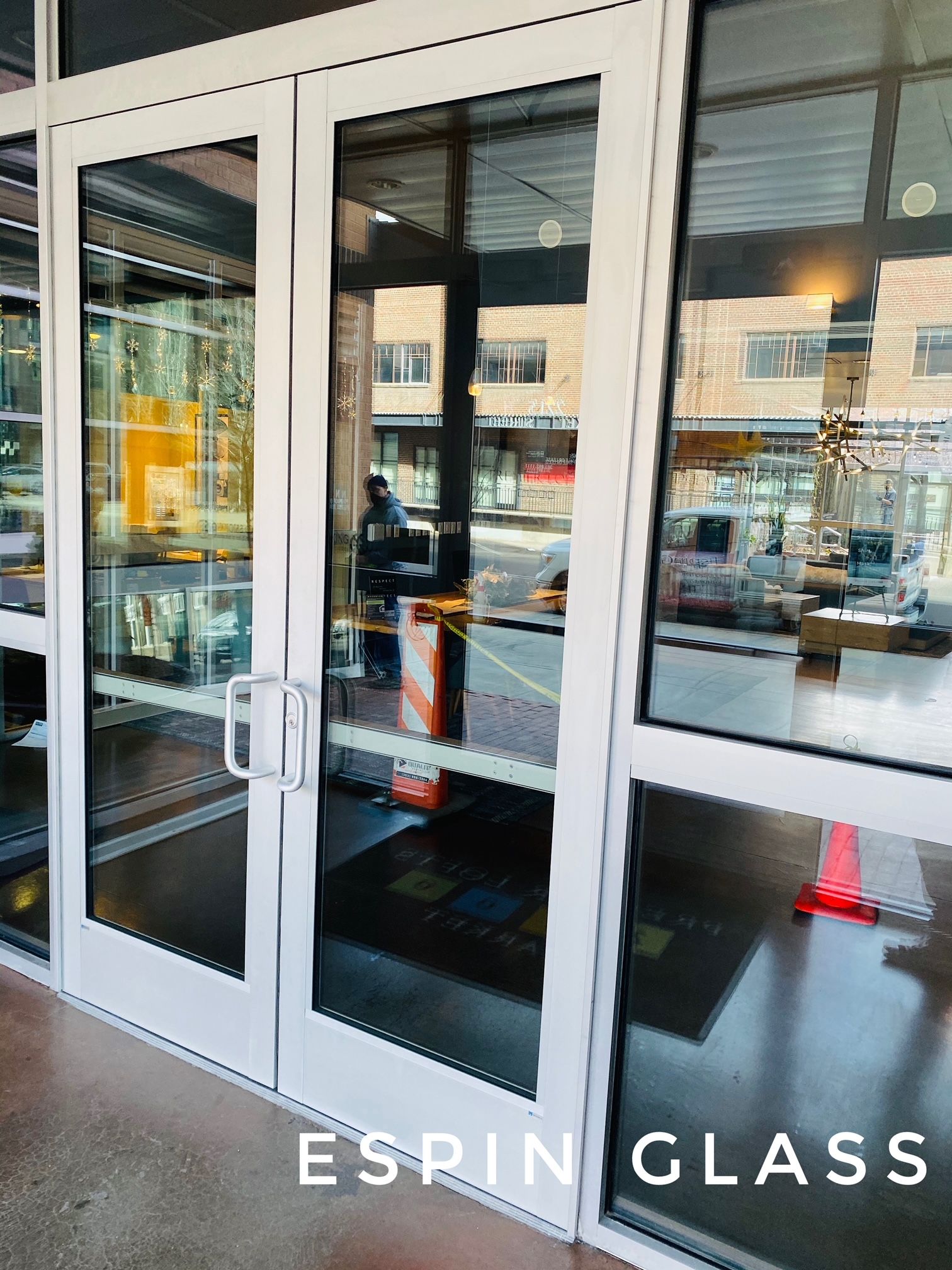 White-framed glass doors reflect a street scene; a business entrance with a white door handle. ESPIN GLASS text at the bottom.