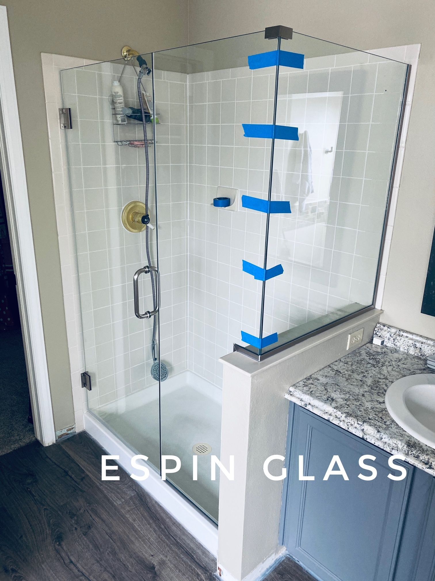 Glass shower enclosure with gold fixtures, installed between white tiled wall and gray vanity.