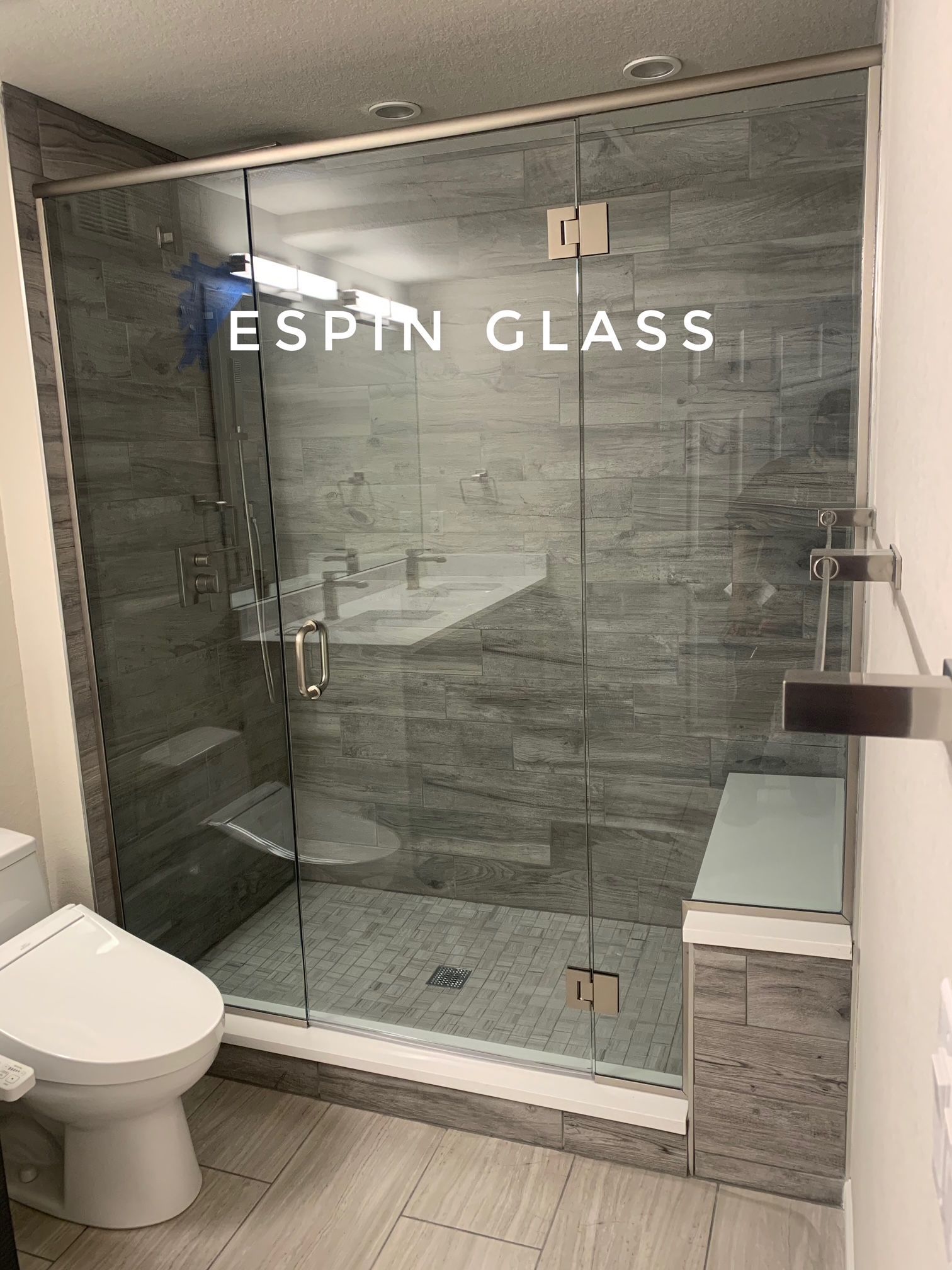 Bathroom with glass shower door, gray tile, toilet, and bench. 
