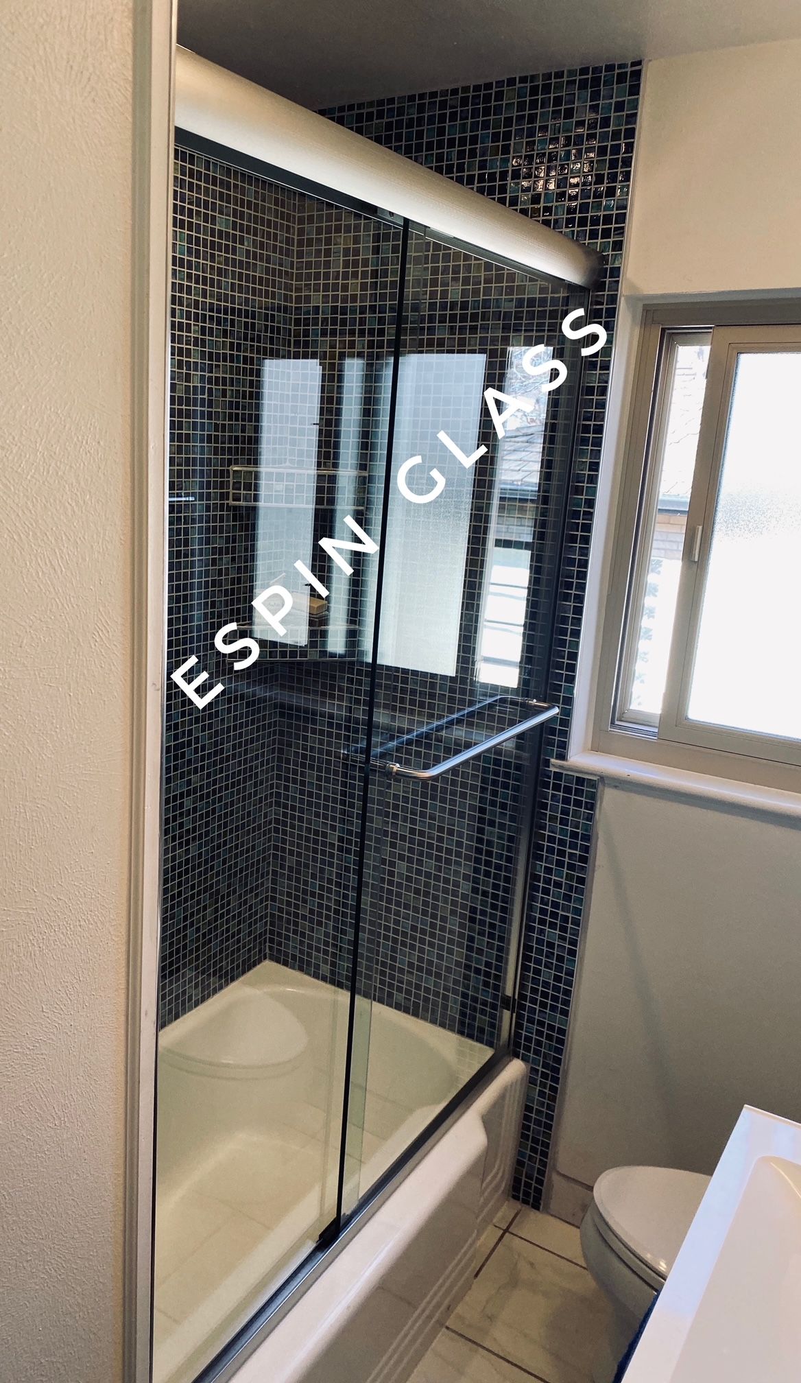 A glass shower door with chrome hardware over a white bathtub. The wall is covered in black and white mosaic tiles.