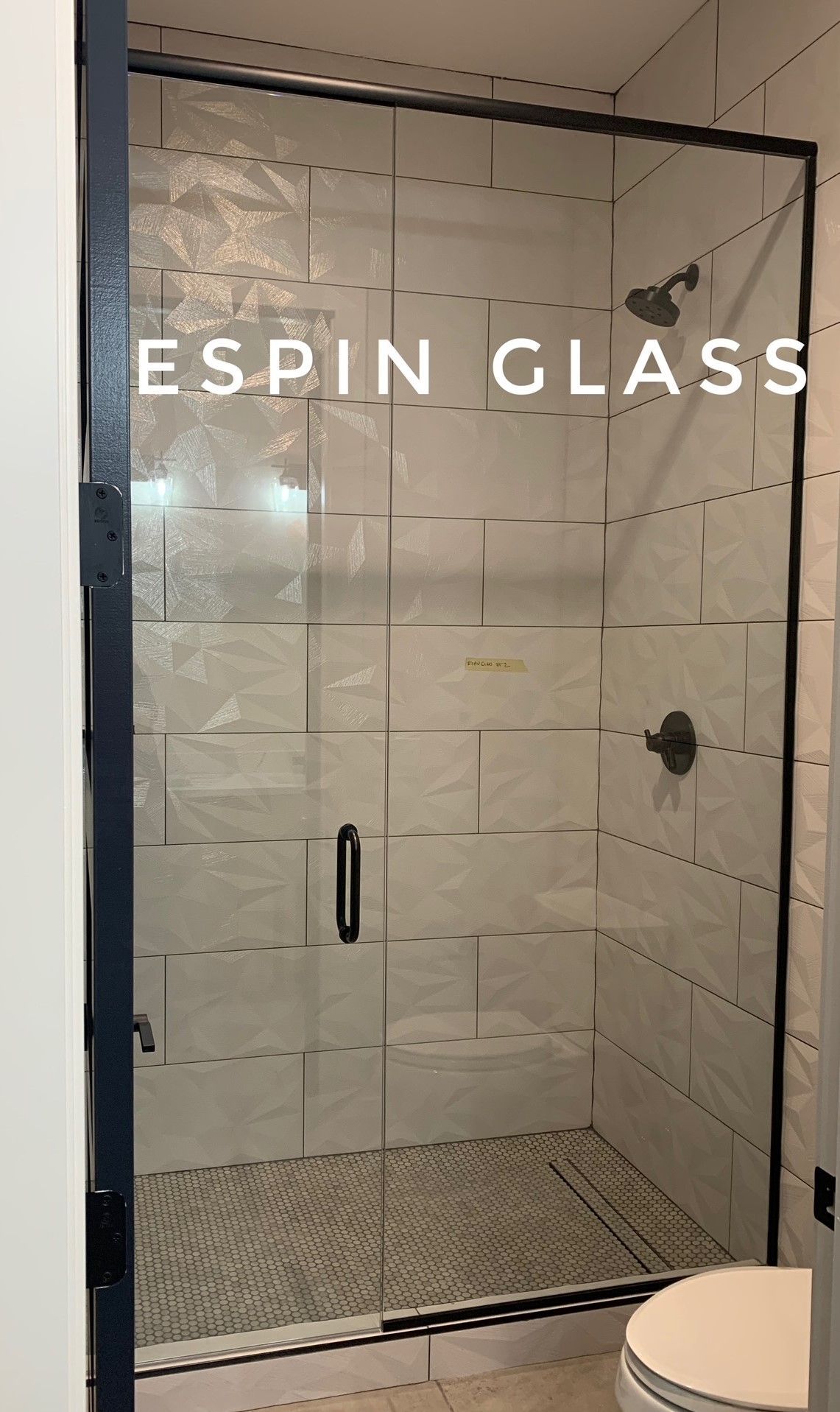 Black-framed glass shower enclosure with white tile walls, bronze fixtures, and a toilet in the corner.