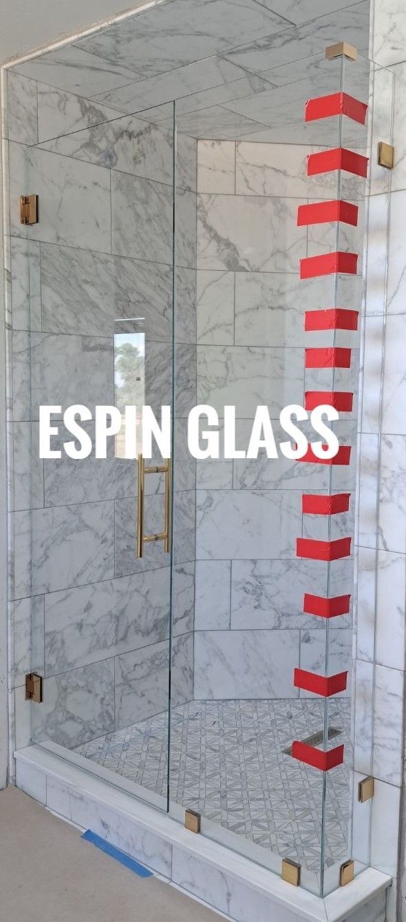 Glass shower door with marble tile walls, held together by gold hardware. The company name 