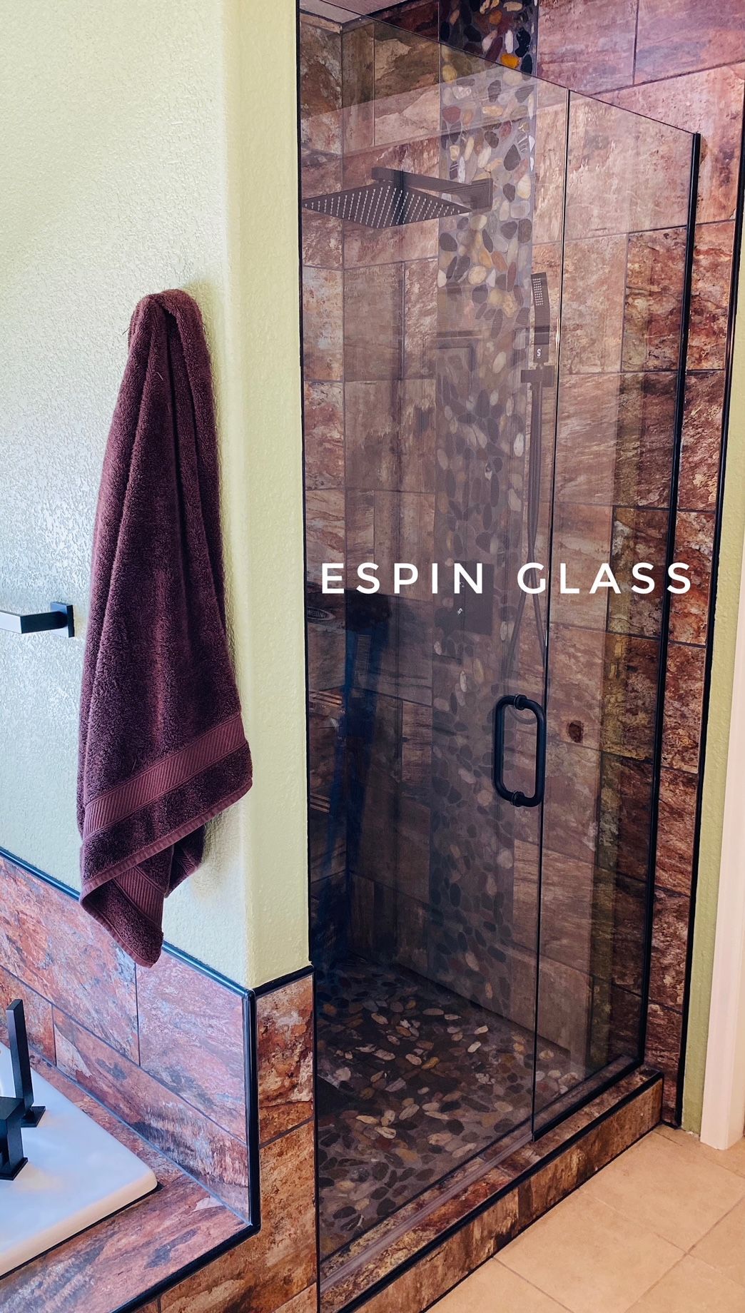 Shower with glass door and stone tile. Burgundy towel on wall.